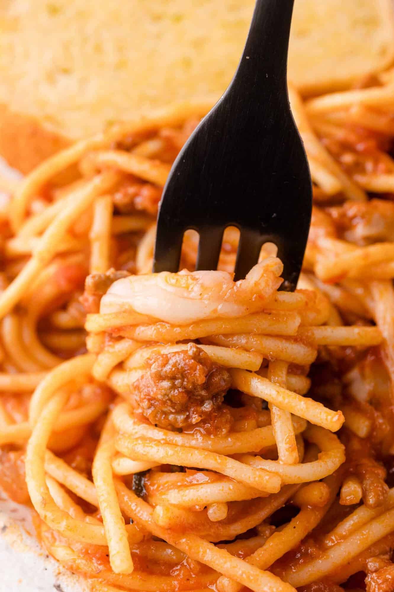 A black fork twirls spaghetti with meat sauce, with a piece of garlic bread visible in the background.