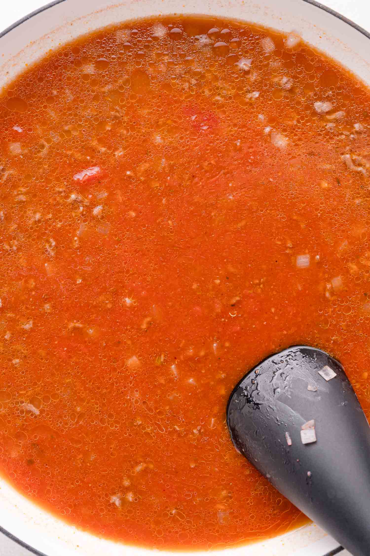 A pot of tomato-based sauce with visible meat and diced onions, being stirred with a black utensil.
