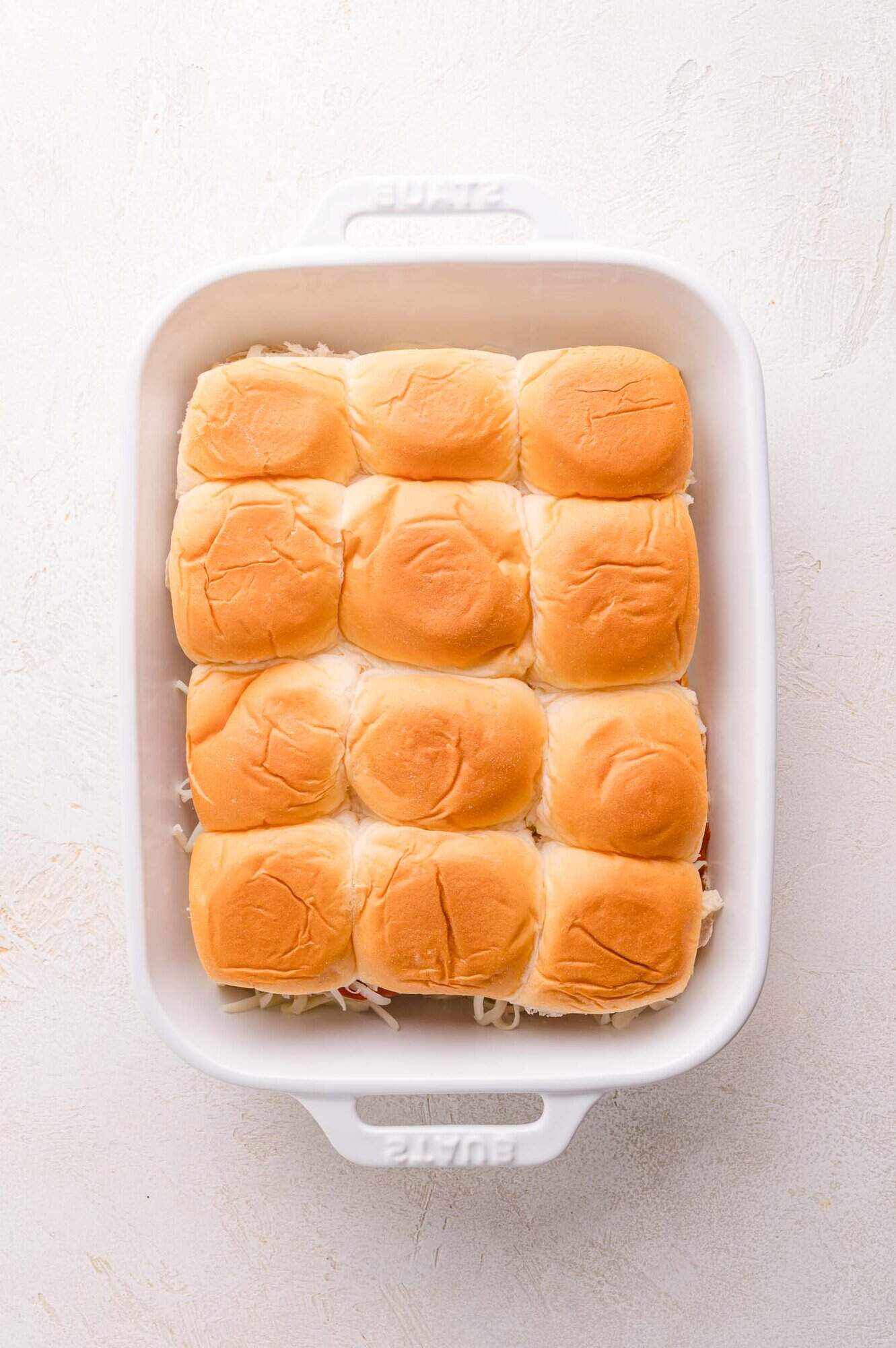 A white baking dish containing a single layer of twelve sandwich rolls arranged in three rows of four.