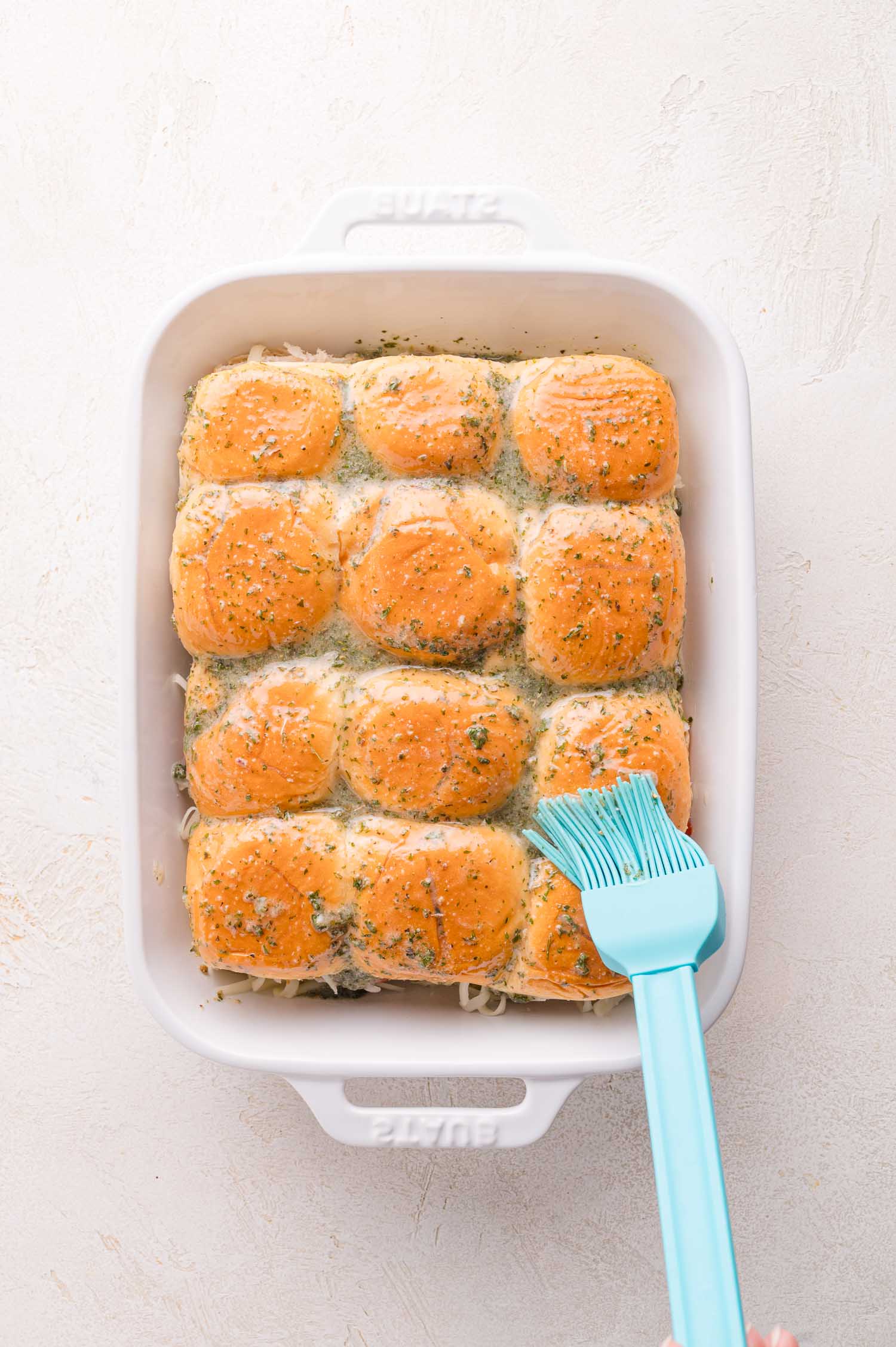 A white baking dish with twelve golden-brown dinner rolls, some topped with herbs, being brushed with melted butter using a blue silicone brush.