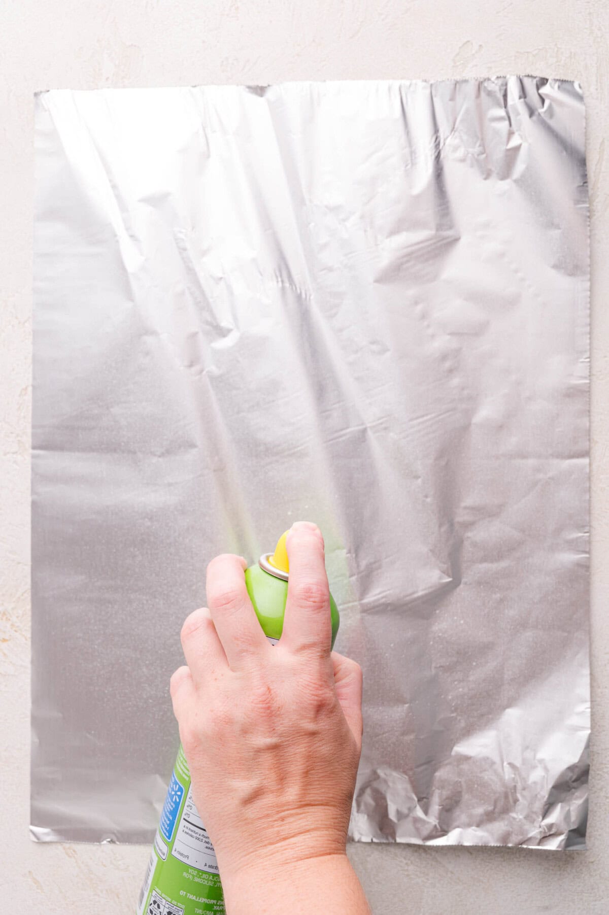 A hand sprays cooking oil from a green can onto a sheet of aluminum foil placed on a light-colored surface.