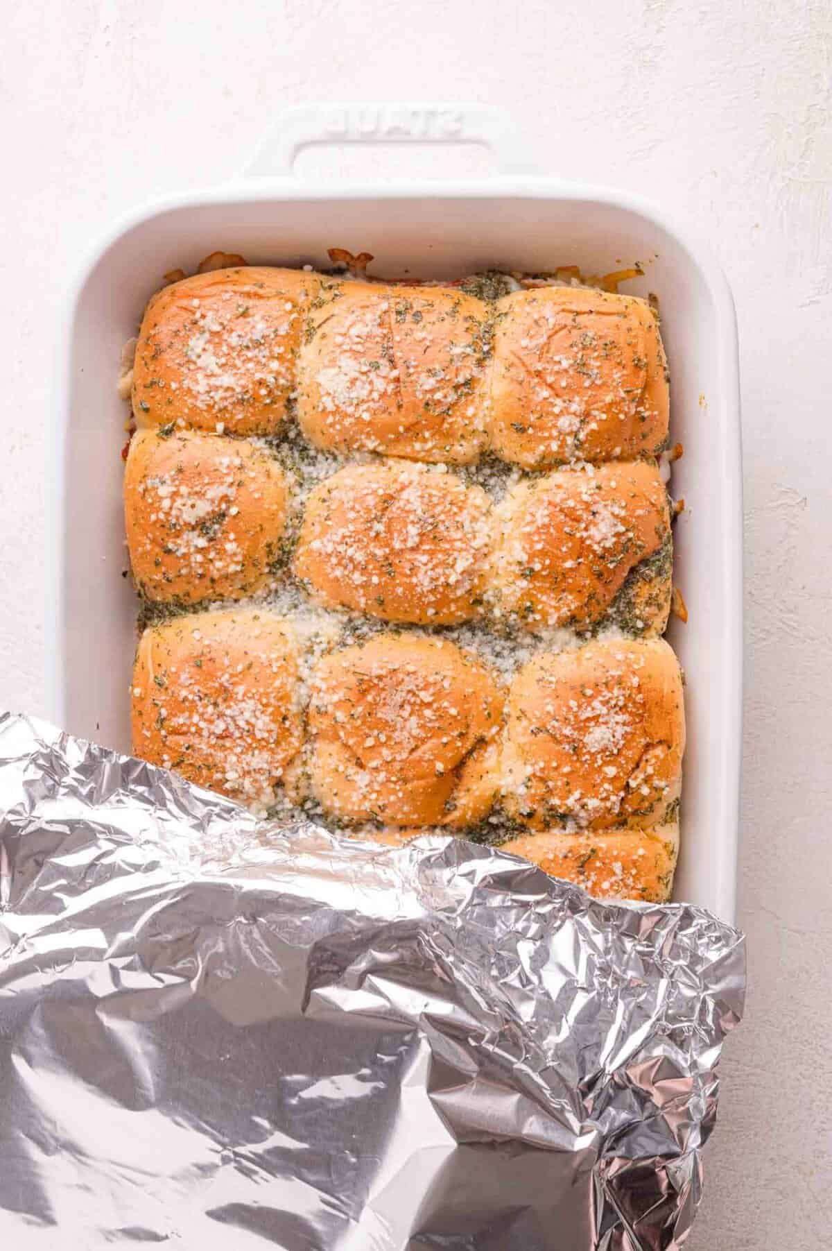 A baking dish filled with golden brown dinner rolls topped with grated cheese and herbs, partially covered with aluminum foil.