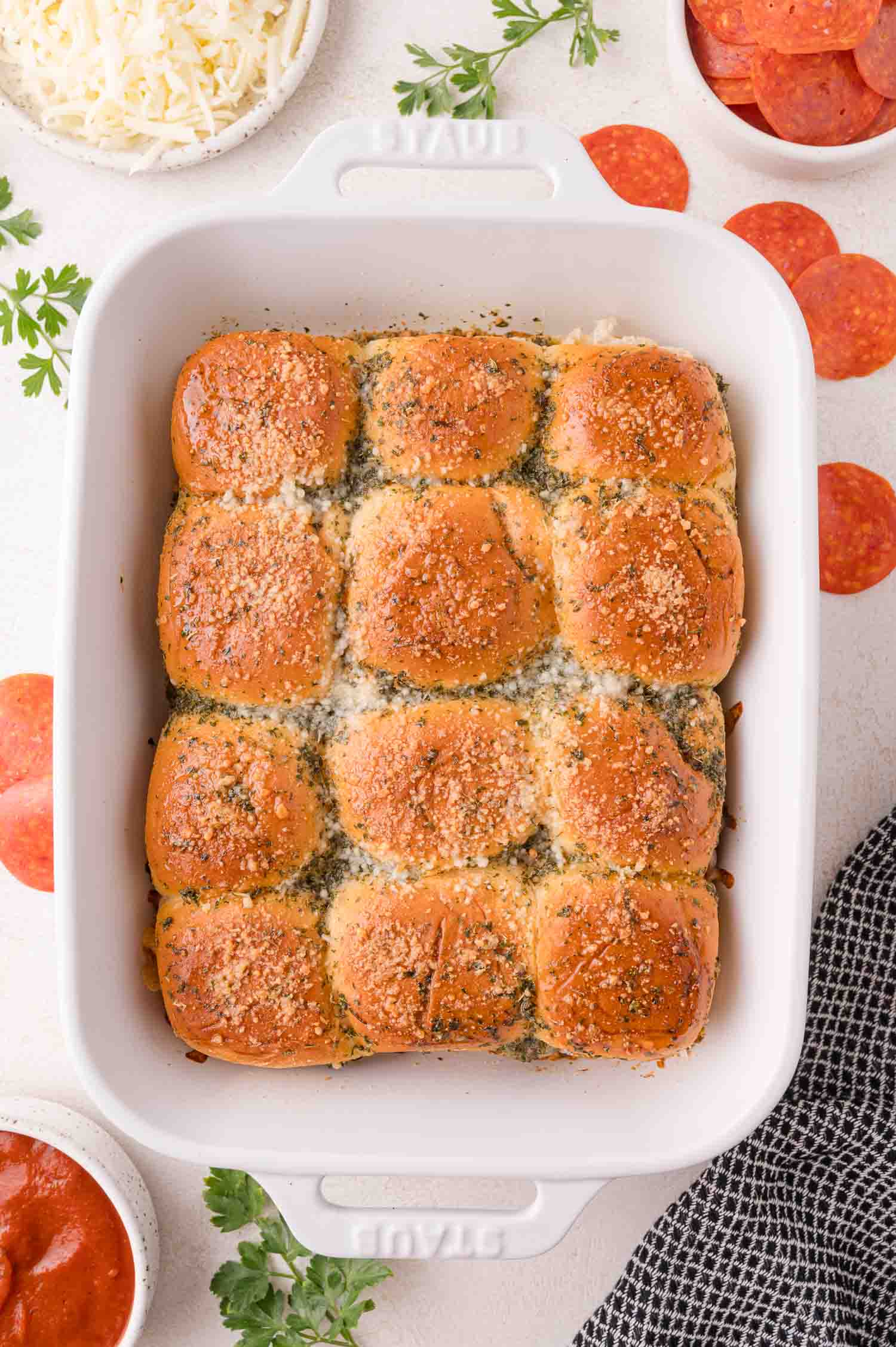 A white baking dish filled with twelve golden-brown slider buns topped with herbs and grated cheese, surrounded by small bowls of shredded cheese, pepperoni, and sauce.