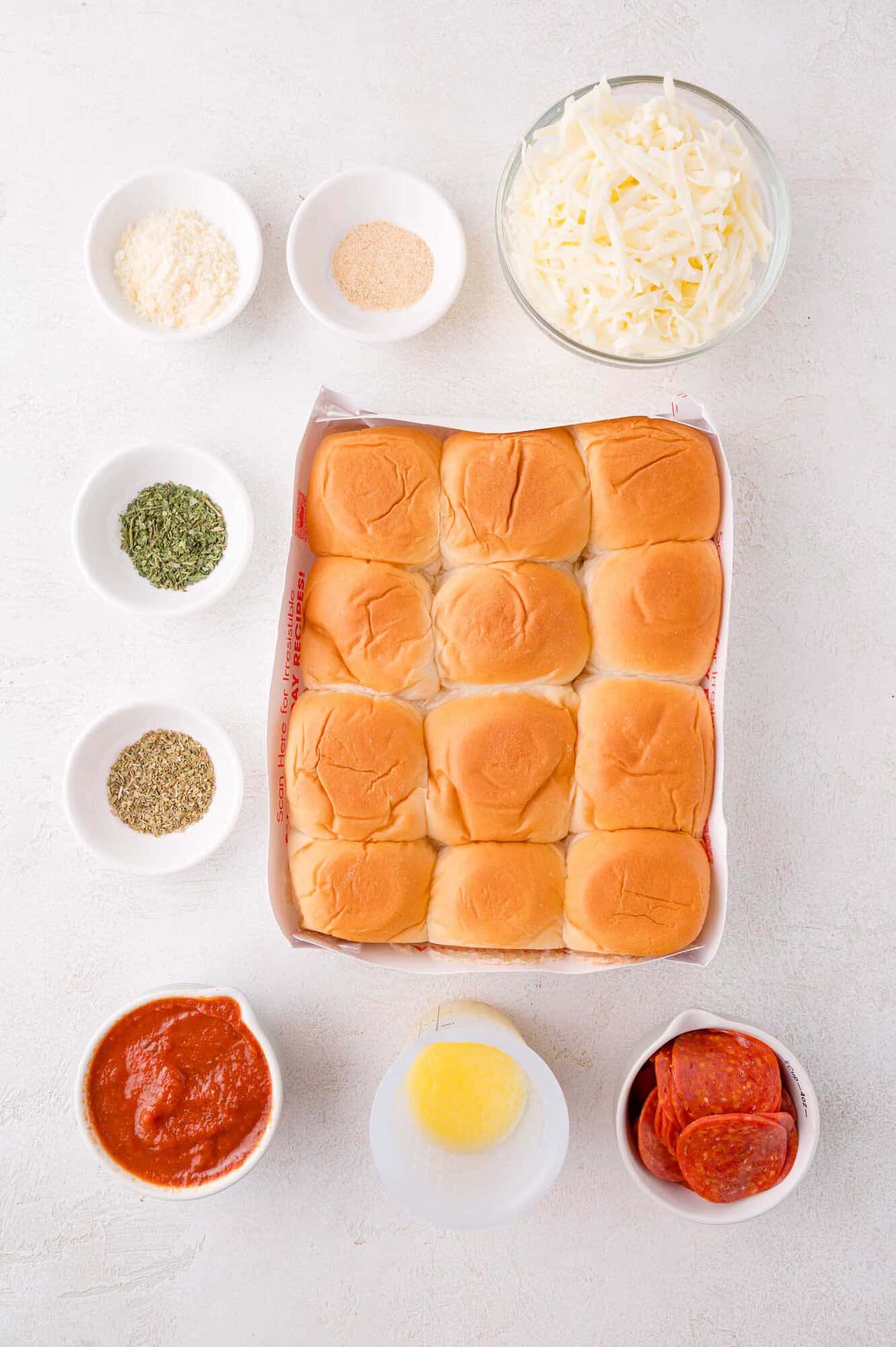 Top-down view of slider buns in a tray surrounded by bowls of shredded cheese, pepperoni slices, marinara sauce, melted butter, and seasonings on a white surface.
