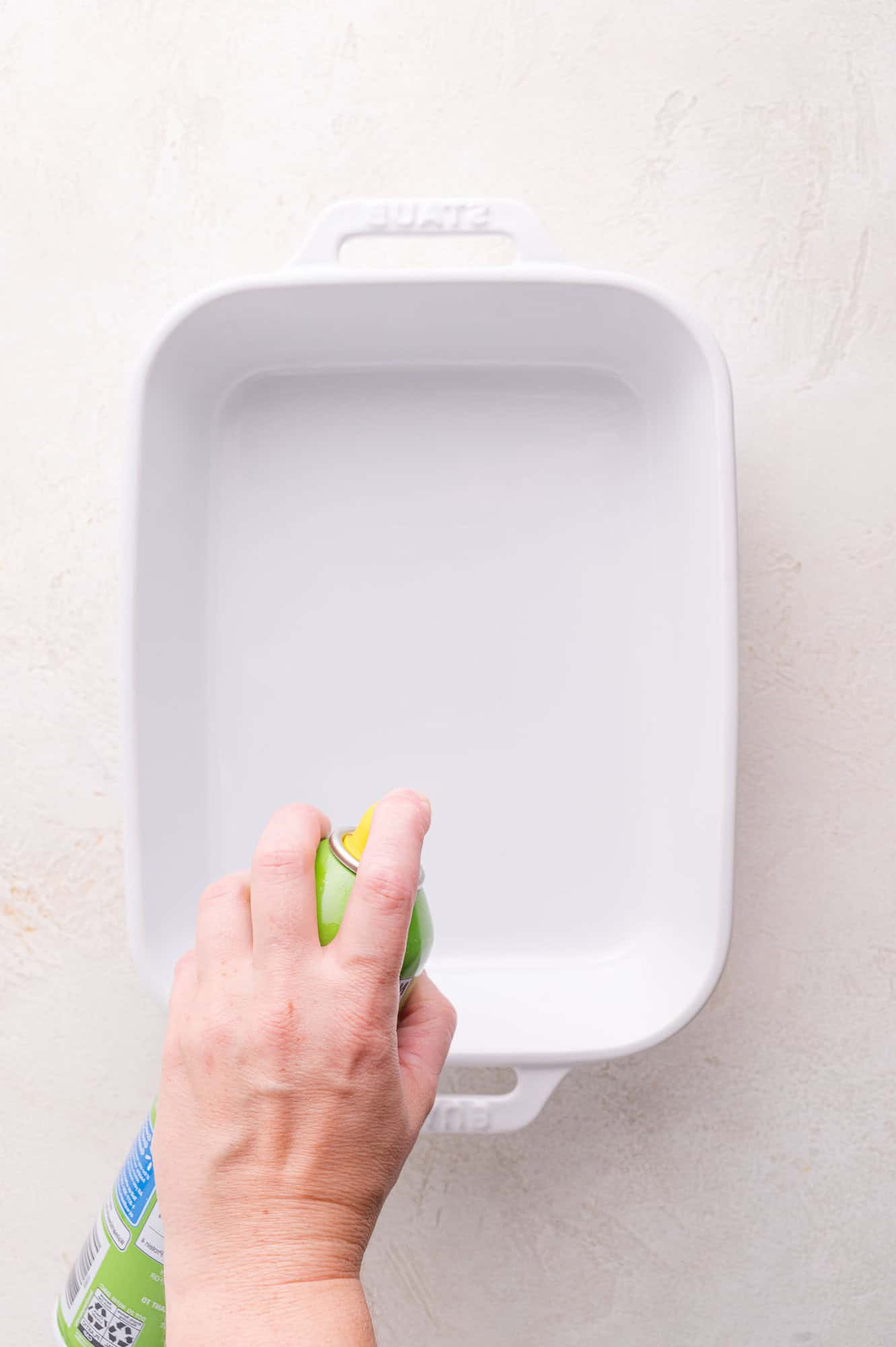 A hand sprays nonstick cooking spray onto a white rectangular baking dish placed on a light-colored surface.