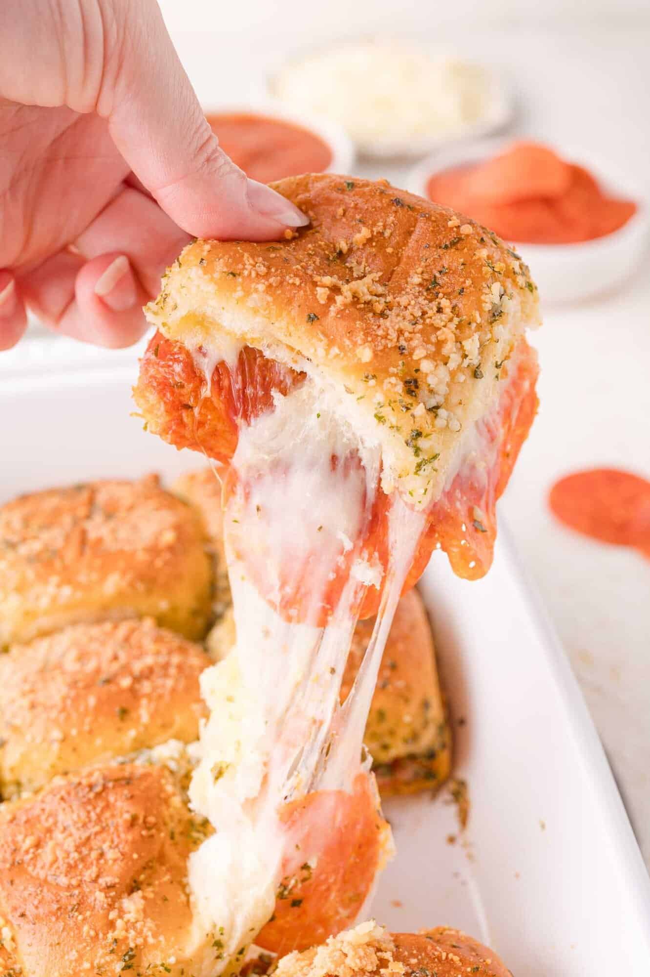 A hand lifts a cheesy pepperoni slider from a baking dish, showing melted cheese stretching from the sandwich.