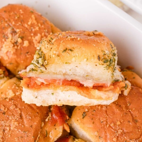 Close-up of a pizza slider sandwich filled with pepperoni and melted cheese, topped with herbs and grated cheese, in a white dish.