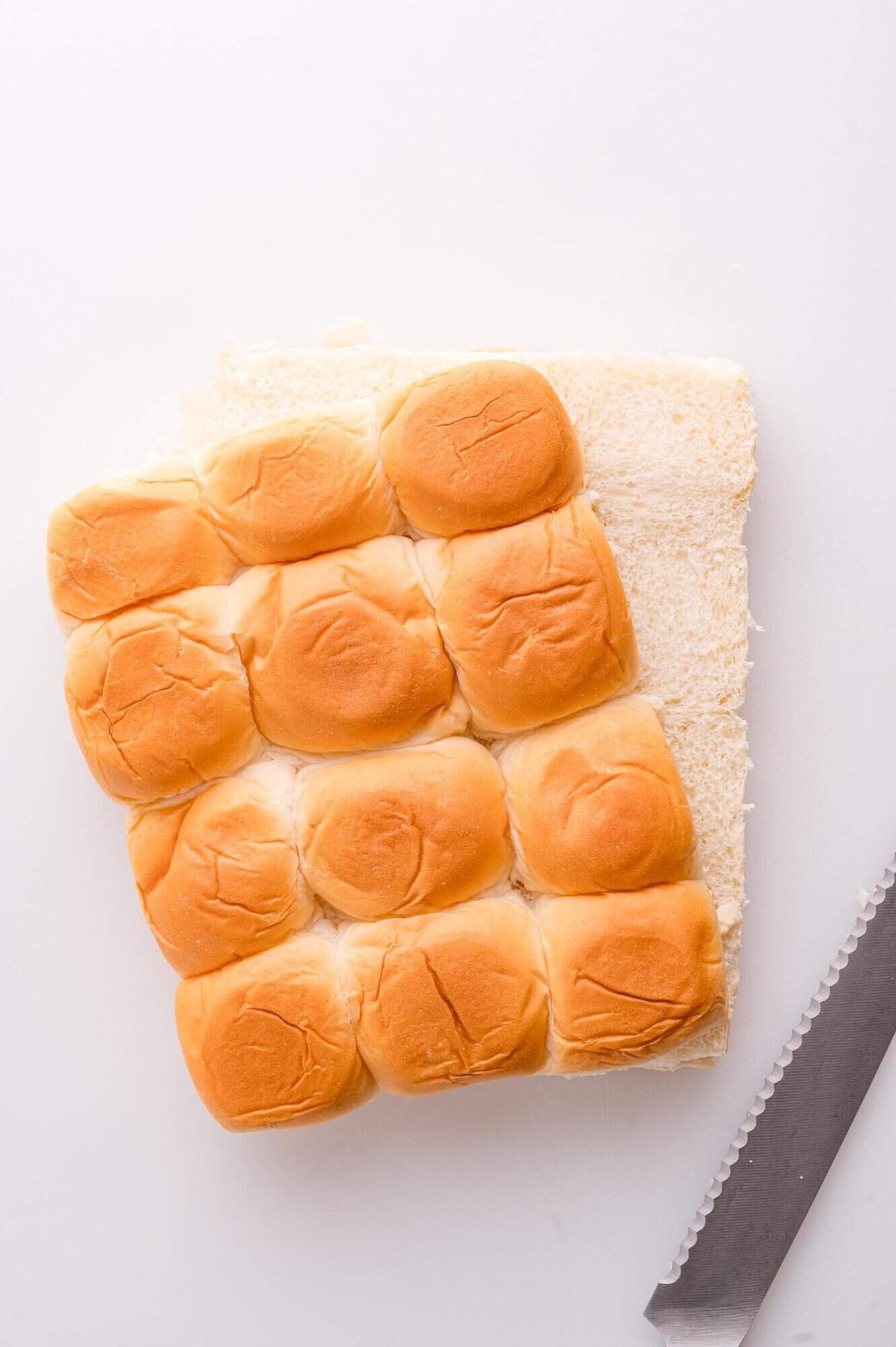 A set of twelve slider buns partially sliced in half on a white surface, next to a serrated knife.