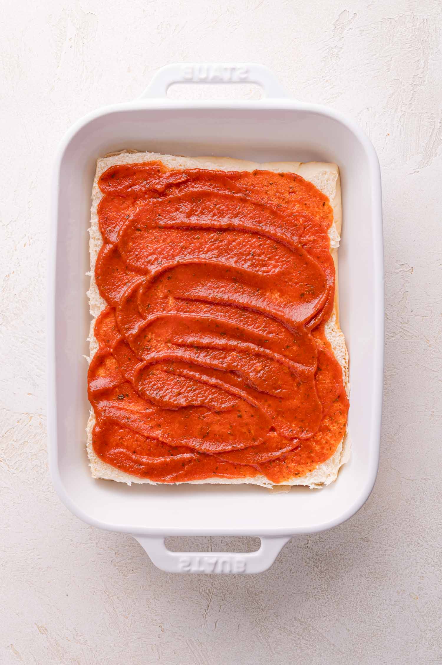A white baking dish with a single layer of lasagna noodles topped with tomato sauce on a light-colored surface.