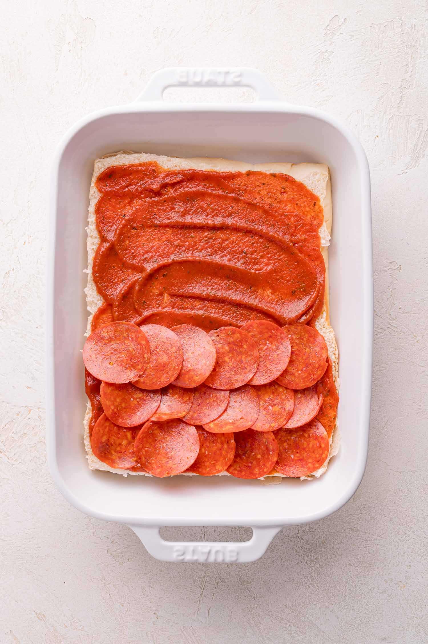 Rectangular white baking dish containing a layer of flat dough, spread with tomato sauce, and topped with slices of pepperoni on one half.