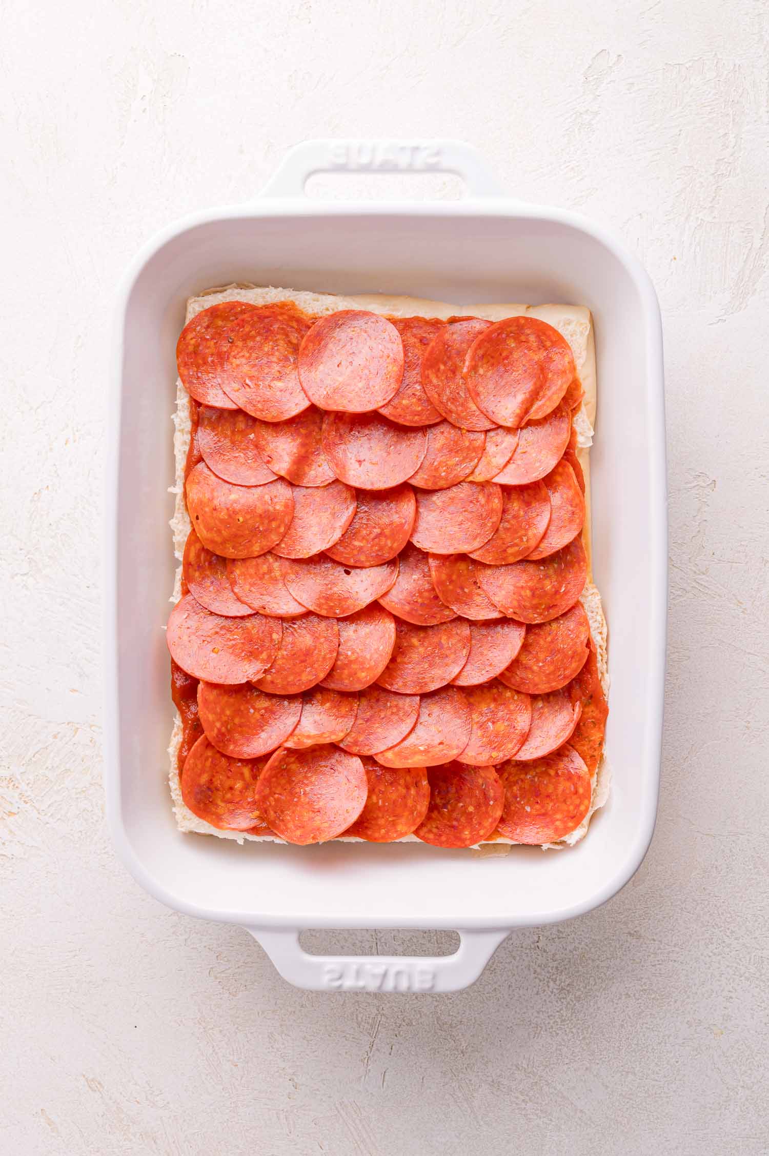 A white baking dish filled with a layer of overlapping pepperoni slices arranged on top of an uncooked rectangular base.