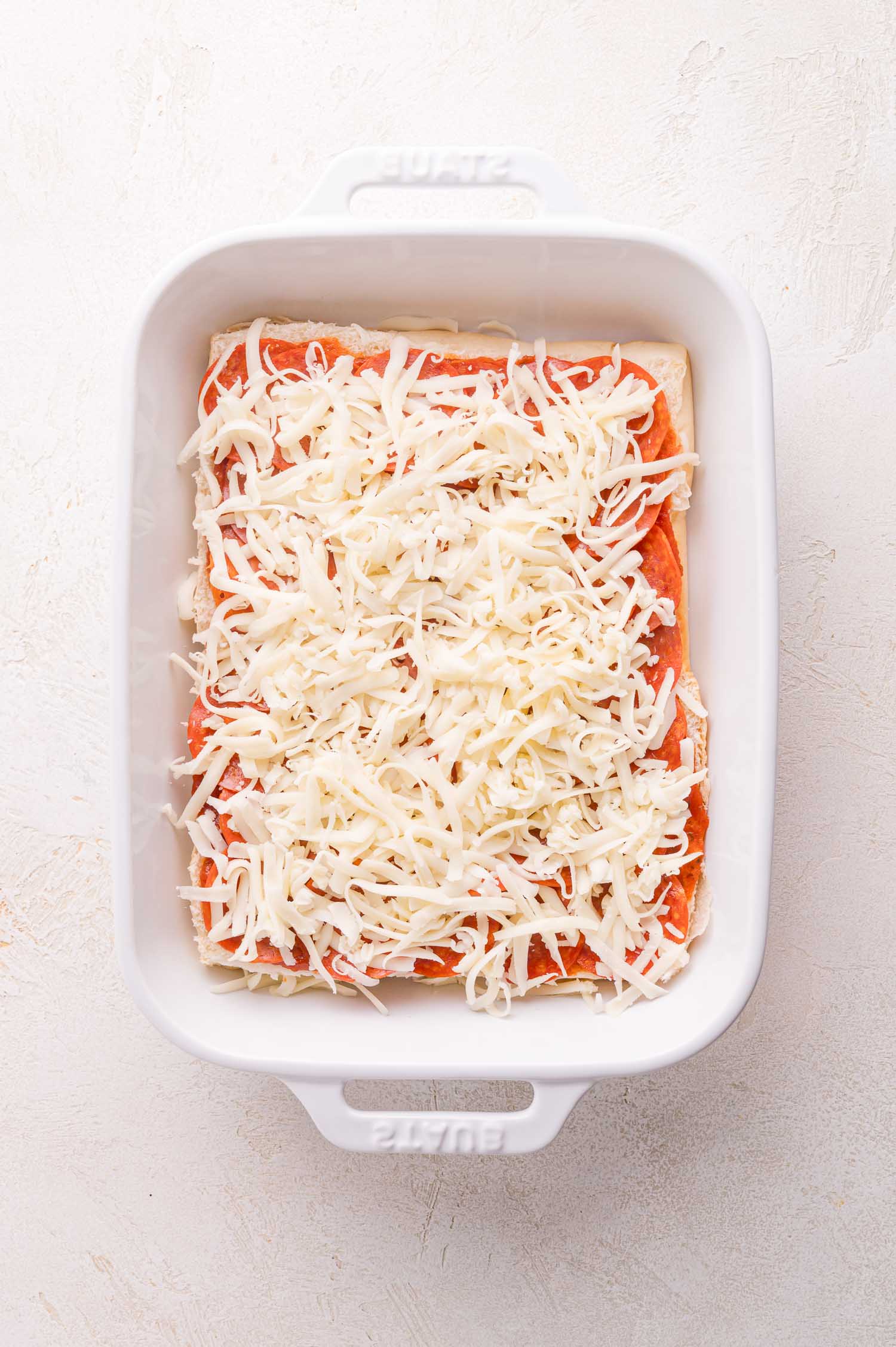 A white baking dish with an unbaked layer of lasagna topped with tomato sauce and shredded cheese.