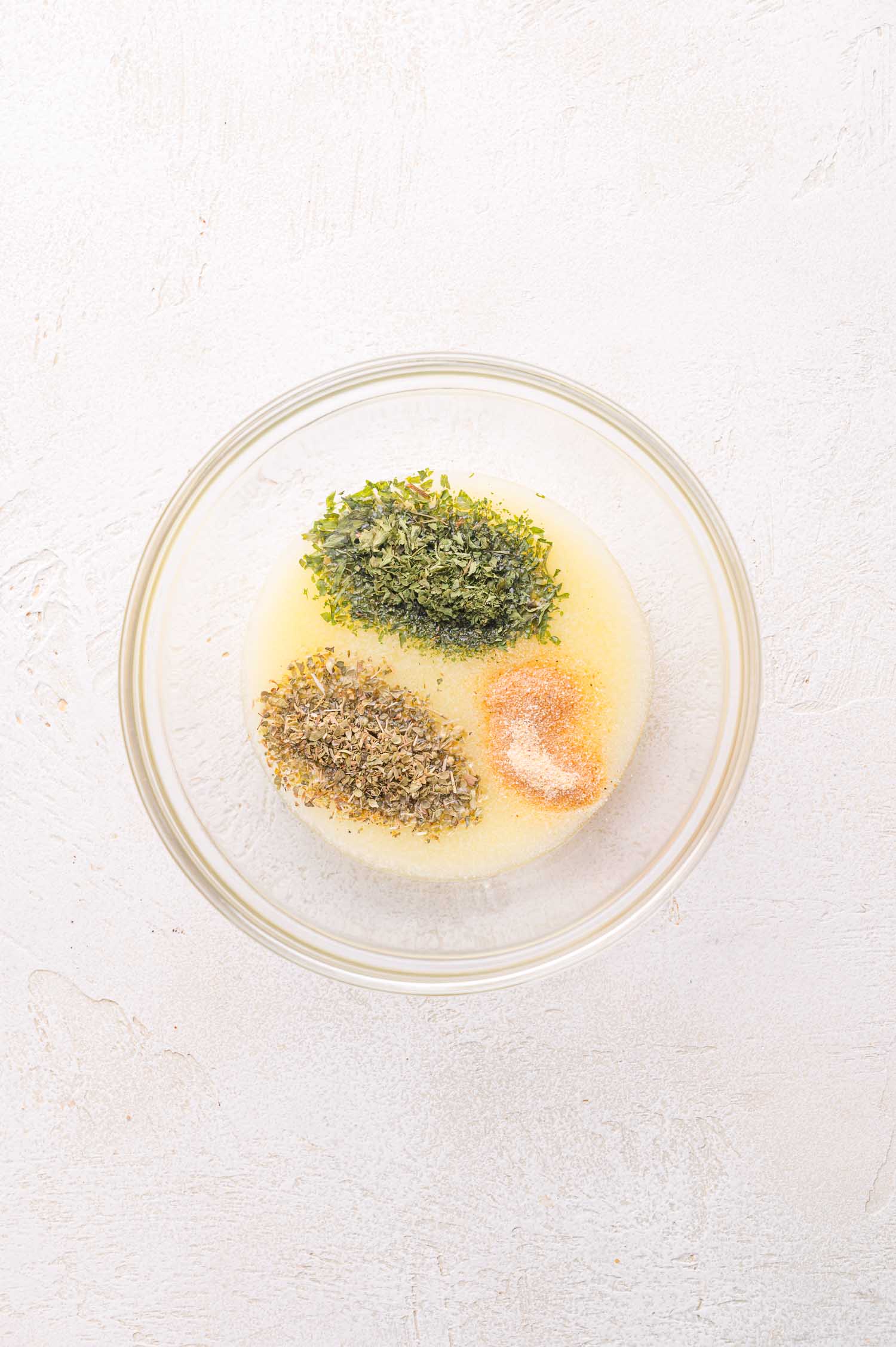 A glass bowl containing melted butter, dried parsley, dried oregano, garlic powder, and salt on a light-colored surface.