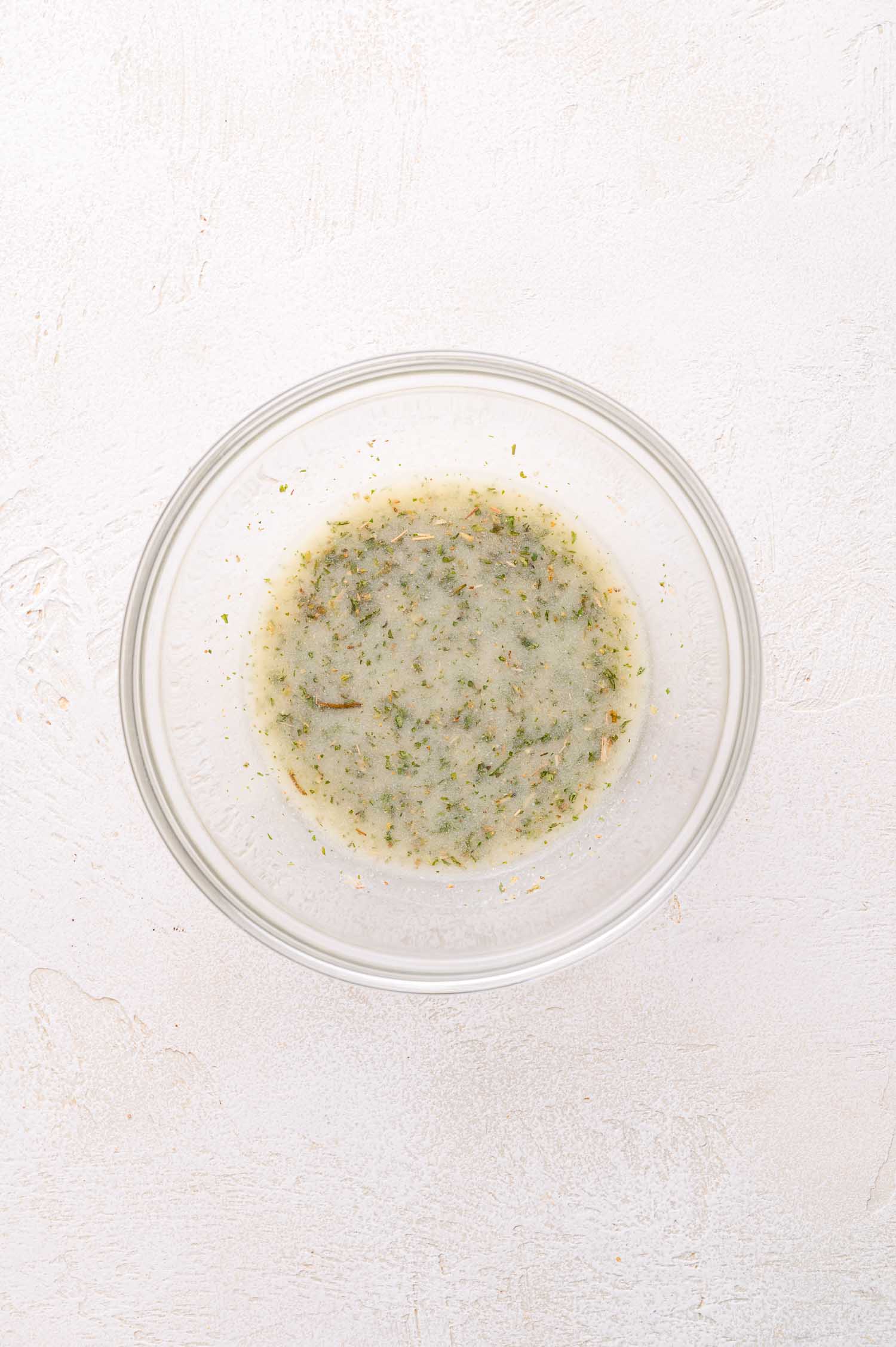 A clear glass bowl containing a light-colored liquid mixed with green and brown dried herbs, placed on a light textured surface.