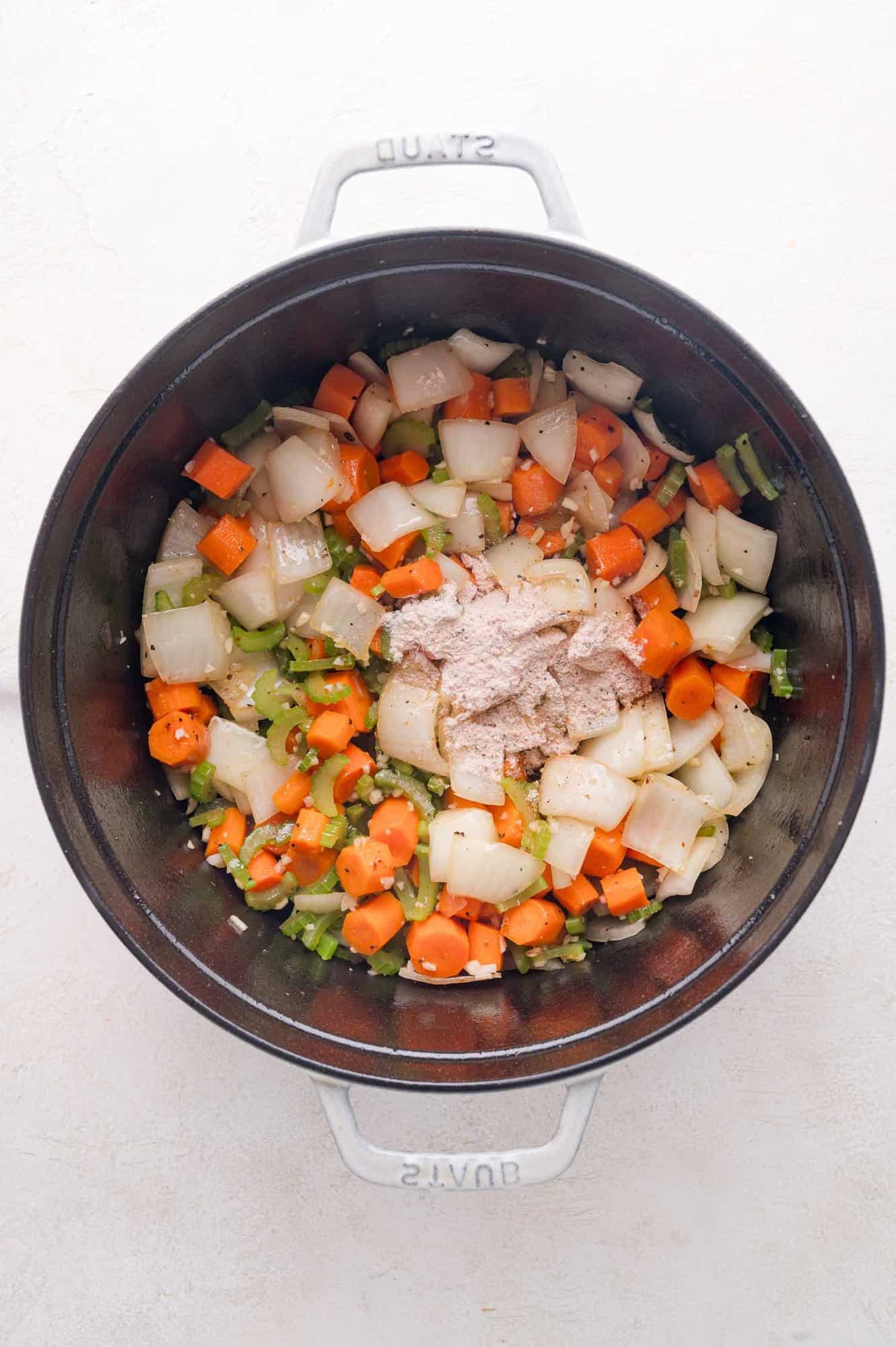 Chopped onions, carrots, and celery sautéing in a black pot, with a mound of seasoned flour added on top.