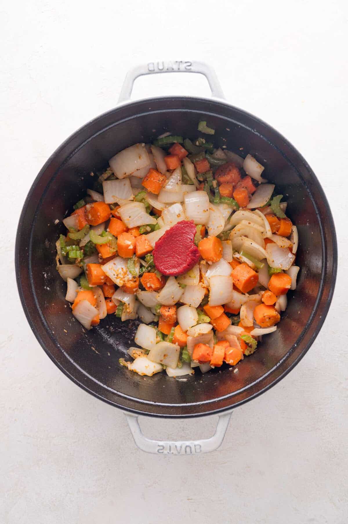 Chopped onions, carrots, and celery with a dollop of tomato paste in a black pot, viewed from above.