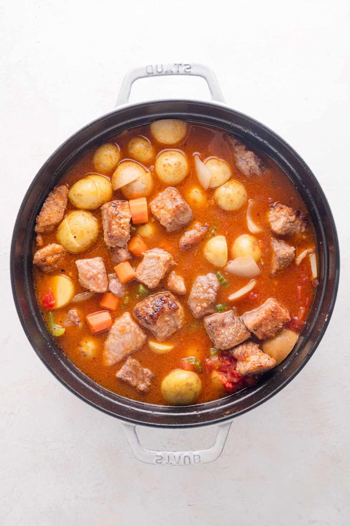 A pot of pork stew with chunks of pork, whole baby potatoes, carrots, onions, and broth, viewed from above on a white background.