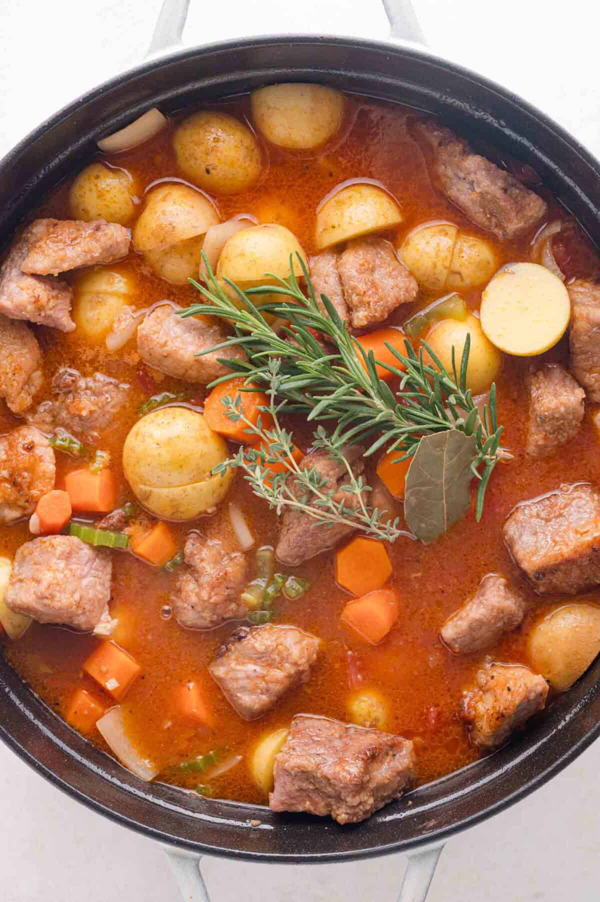 A pot of pork stew with chunks of pork, baby potatoes, carrots, and herbs in a rich broth, garnished with sprigs of rosemary and bay leaf.