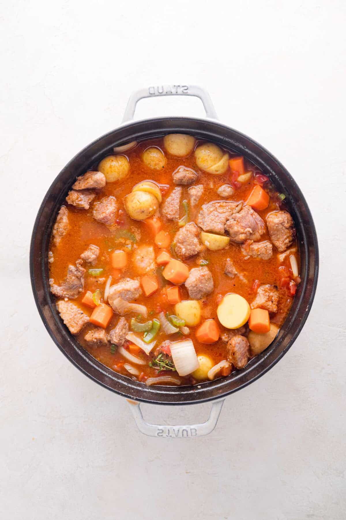 A pot of stew with chunks of pork, potatoes, carrots, onions, and celery in a red broth.