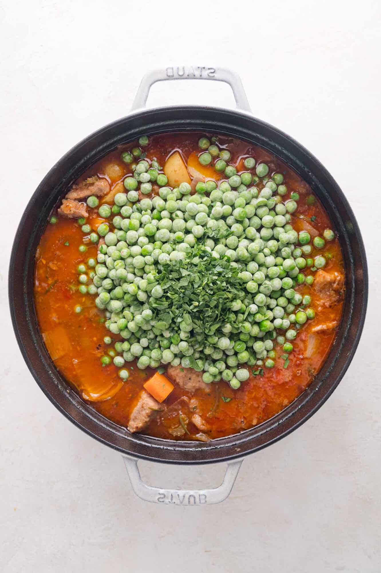 A pot of stew containing pork, potatoes, carrots, and tomato sauce, topped with a pile of frozen green peas and chopped parsley.