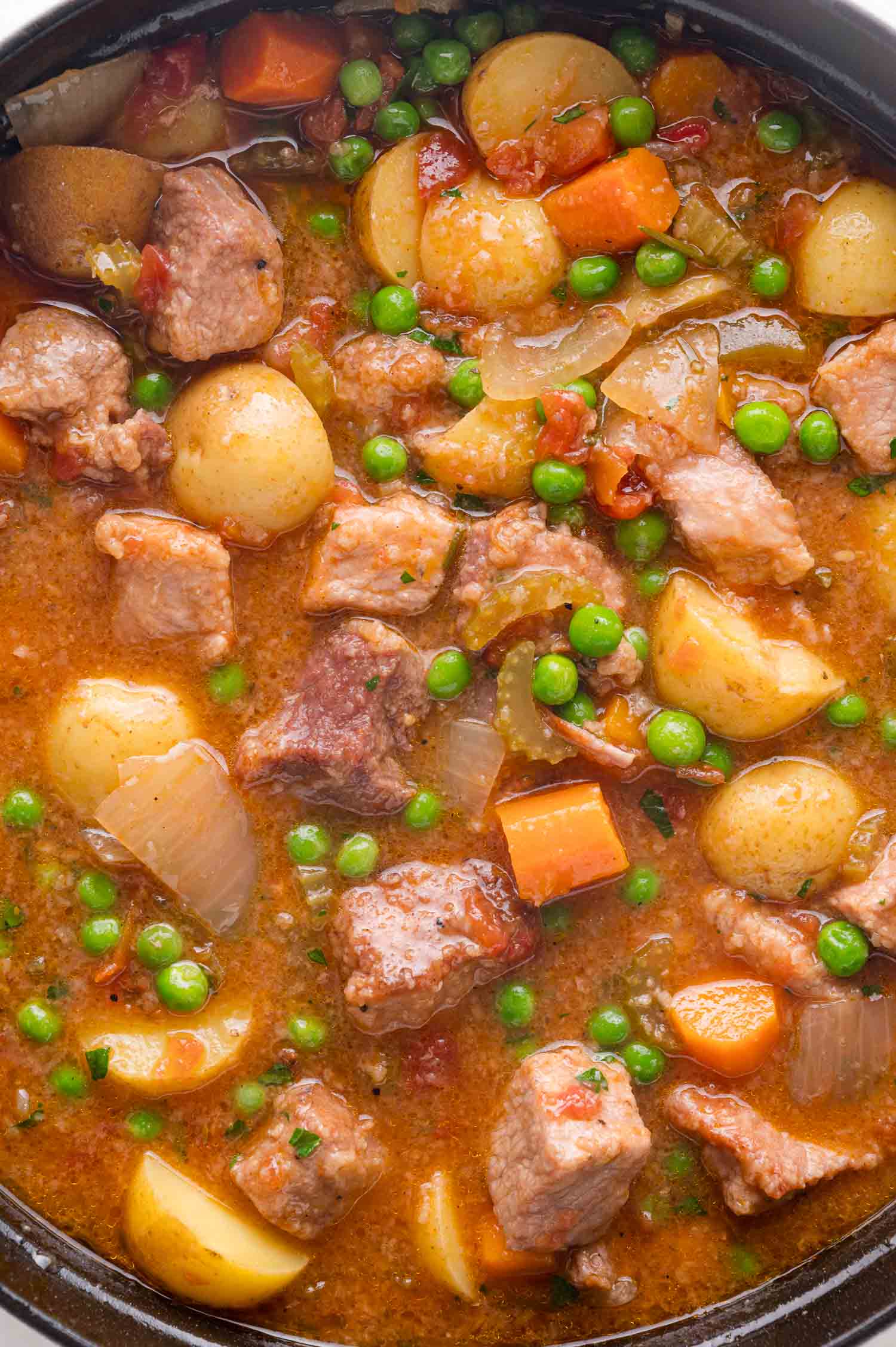 A close-up of a pot filled with pork stew containing chunks of pork, potatoes, carrots, peas, onions, and a rich broth.
