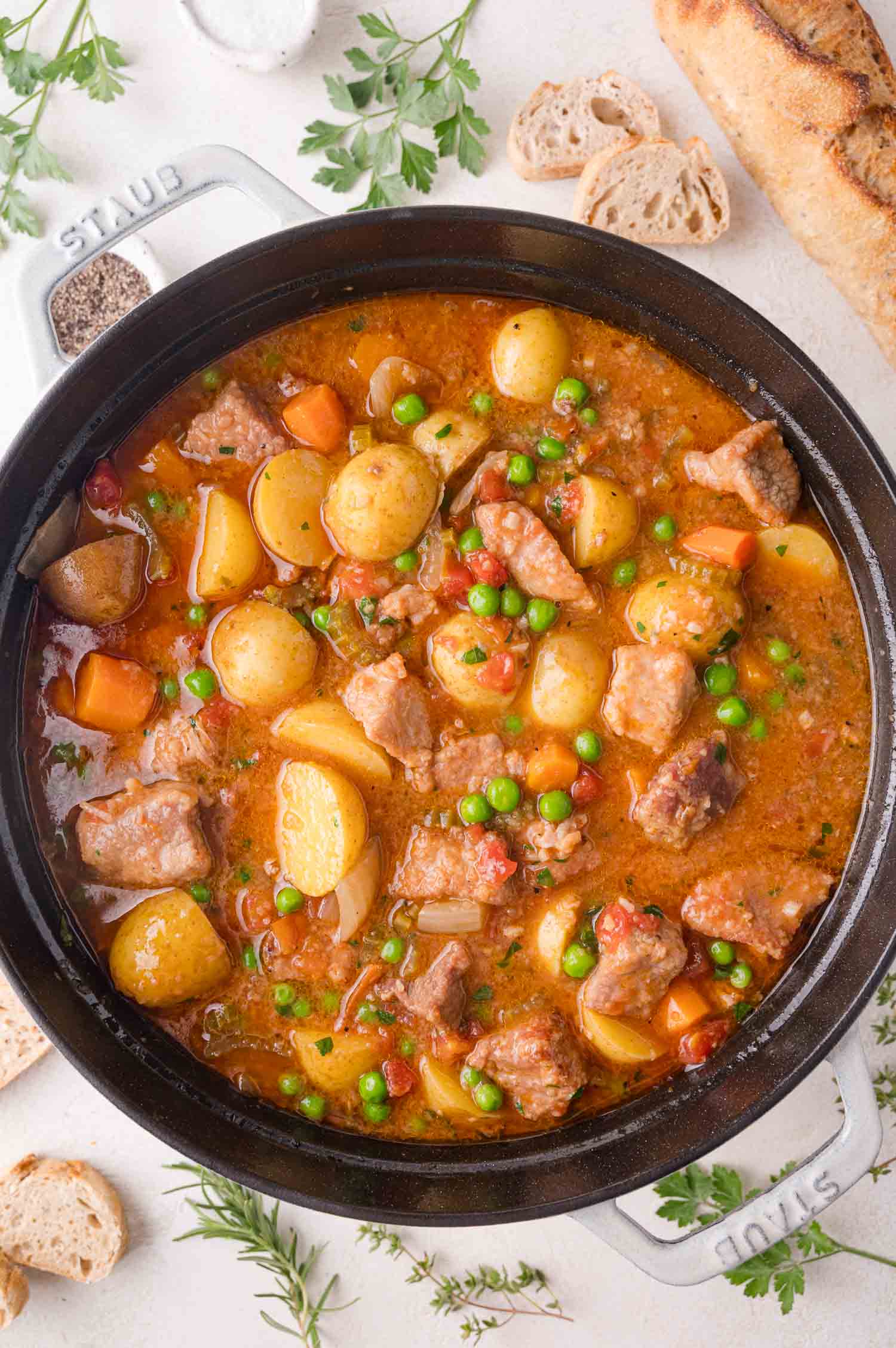 A pot of pork stew with chunks of pork, potatoes, carrots, peas, and a tomato-based broth, surrounded by bread and fresh herbs.