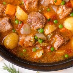 Close-up of a hearty pork stew with chunks of pork, potatoes, carrots, peas, onions, and a rich broth in a black pot.