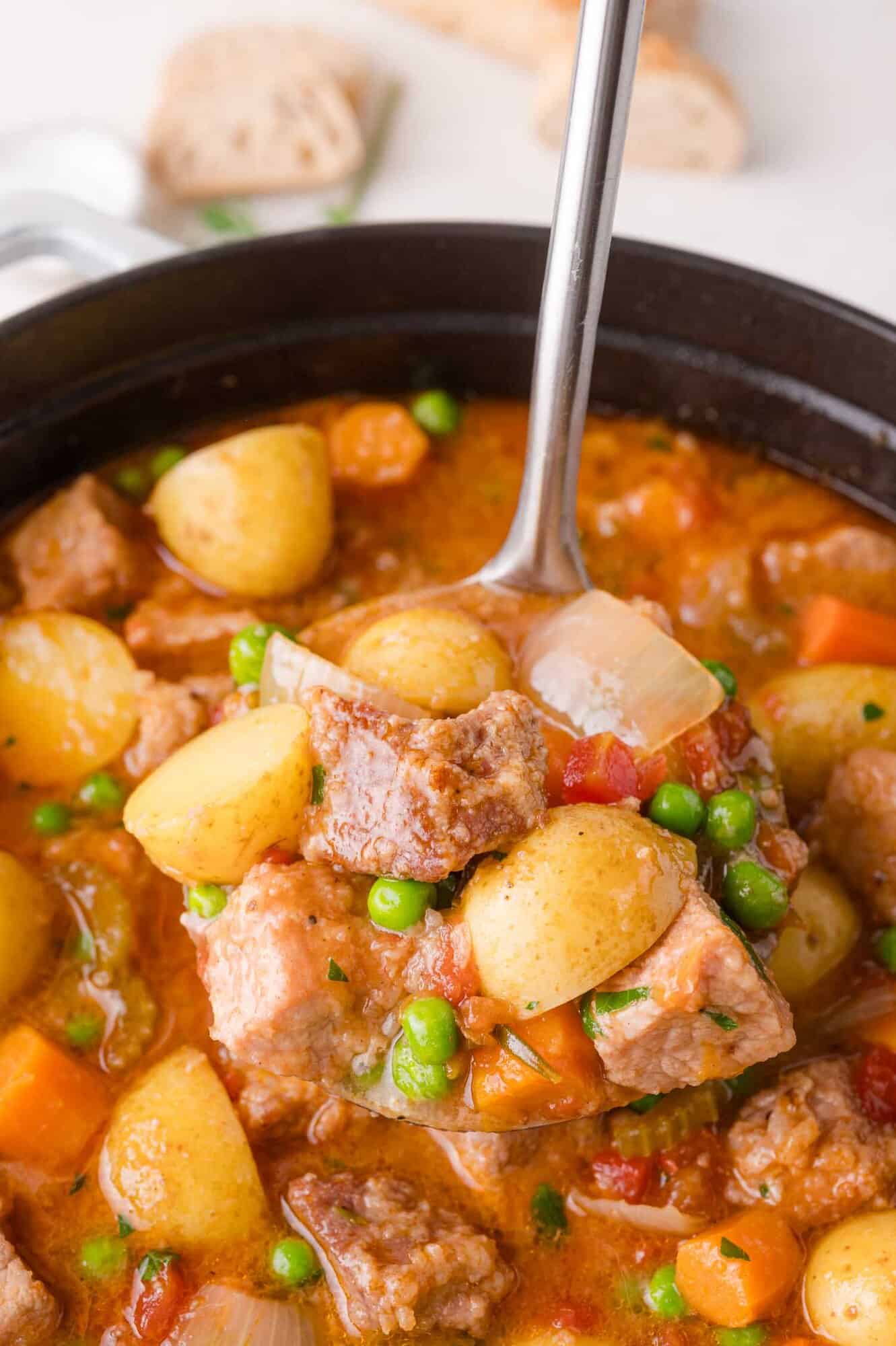 A ladle scoops up pork stew with potatoes, peas, carrots, and onions from a pot.