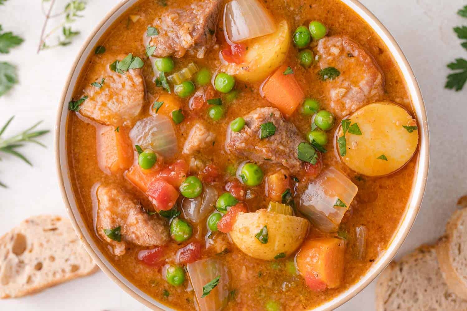 A bowl of hearty stew with pork, potatoes, carrots, peas, onions, celery, and herbs in a rich broth, served next to slices of bread.