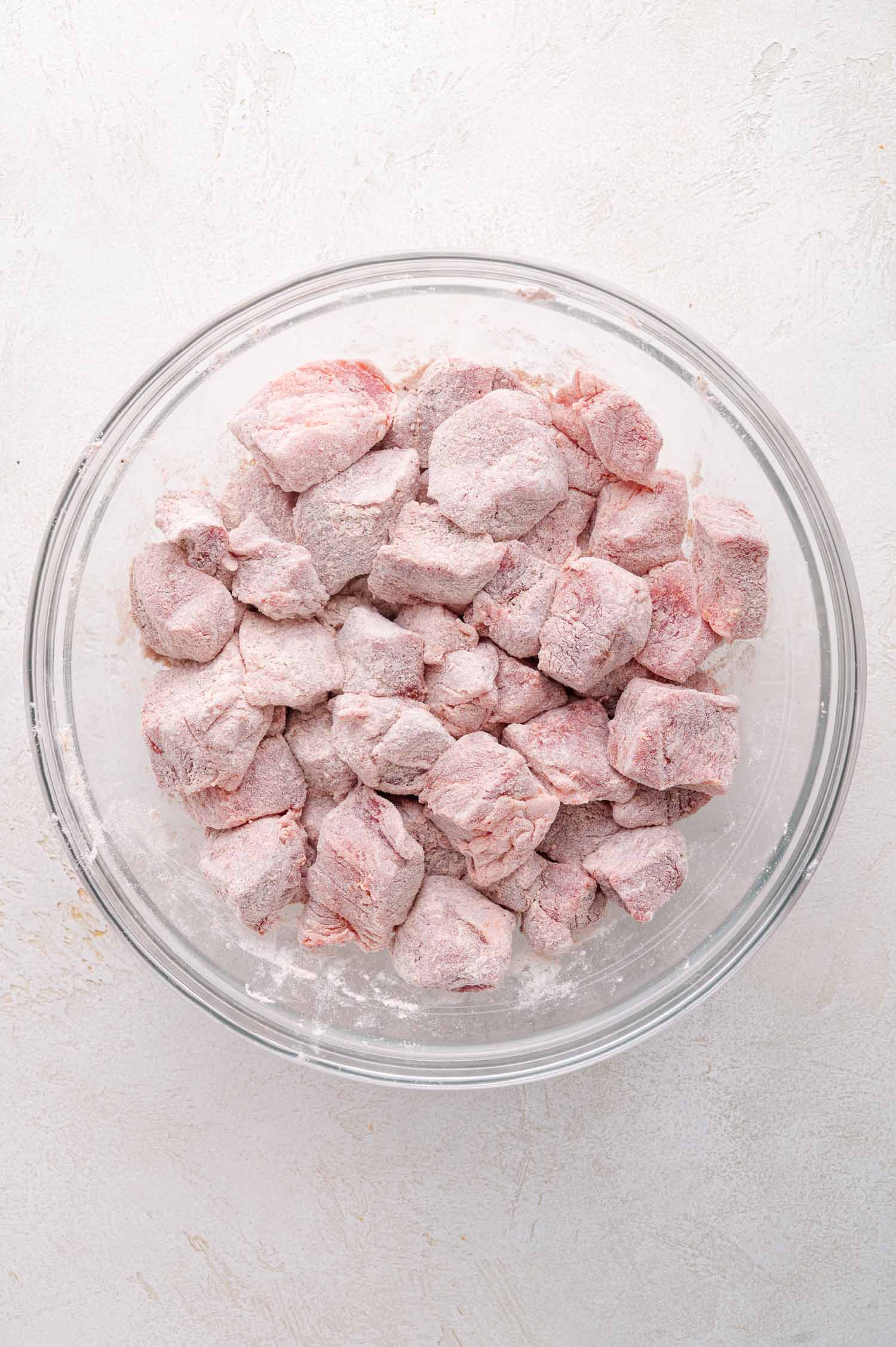 A glass bowl filled with raw pork cubes coated in flour on a light-colored surface.