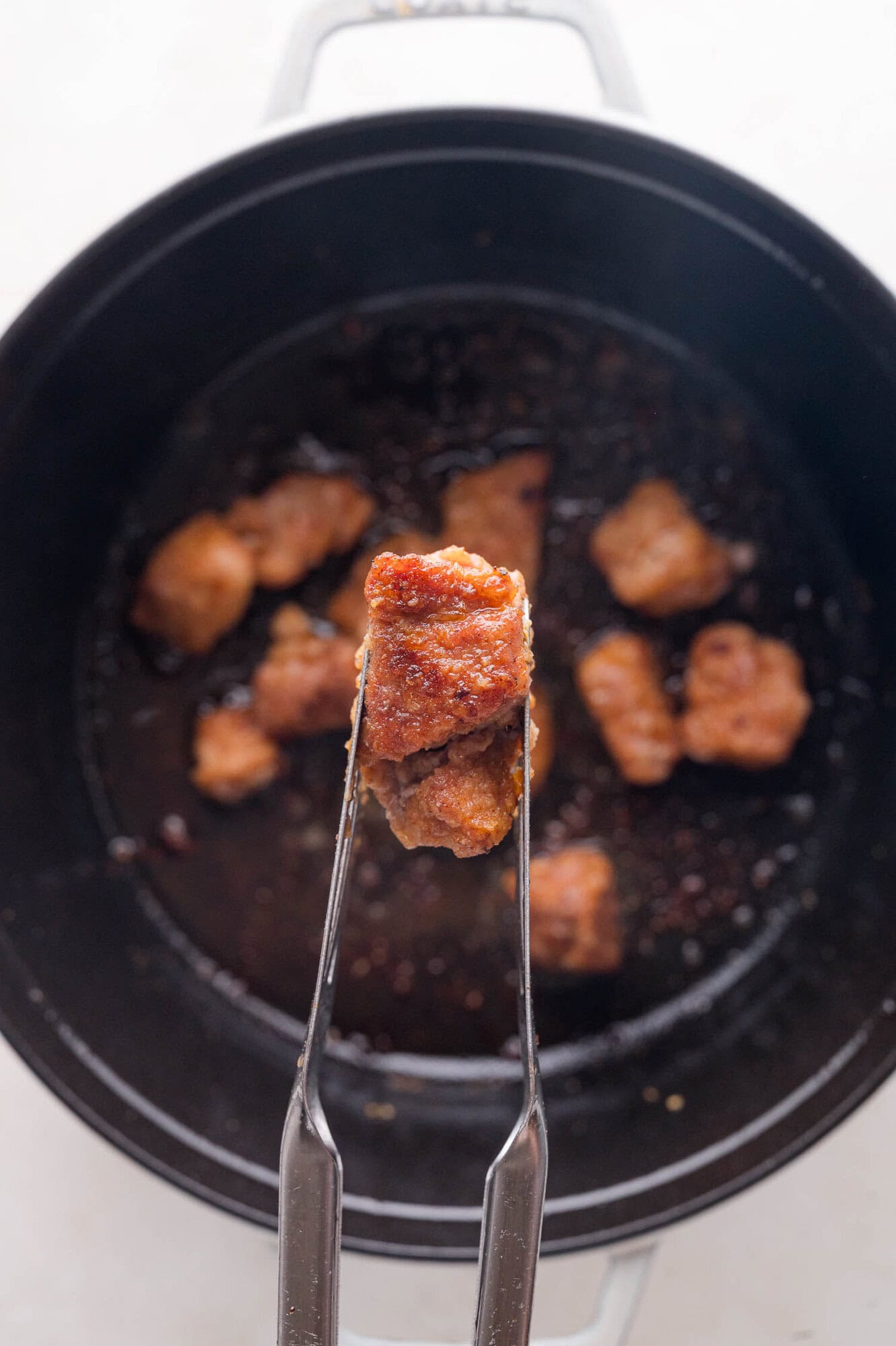 A pair of metal tongs holding a piece of cooked, browned pork above a black pot with more pieces of meat inside.