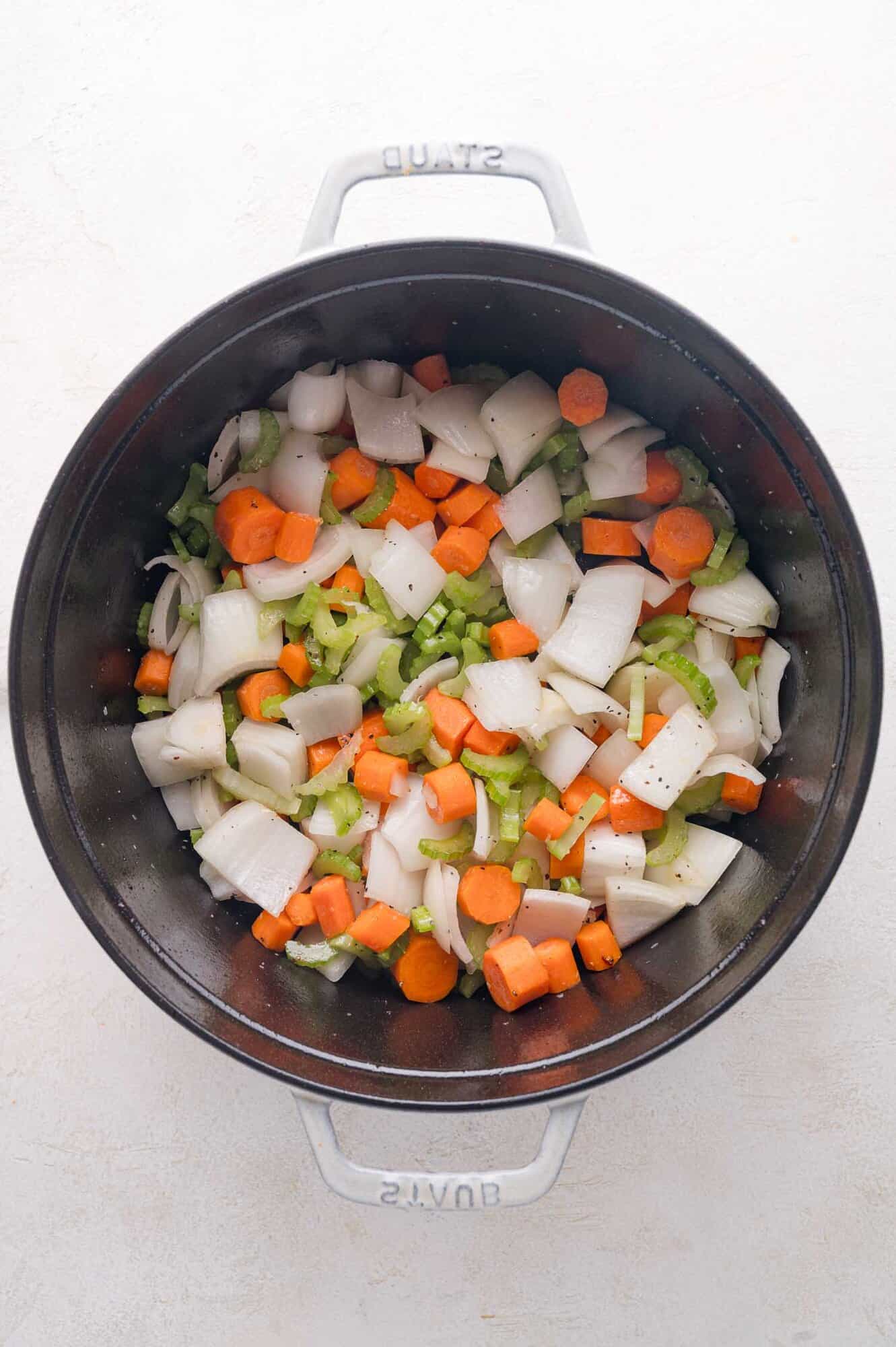 Chopped onions, carrots, and celery are seasoned with salt and pepper in a black Dutch oven, ready for cooking.