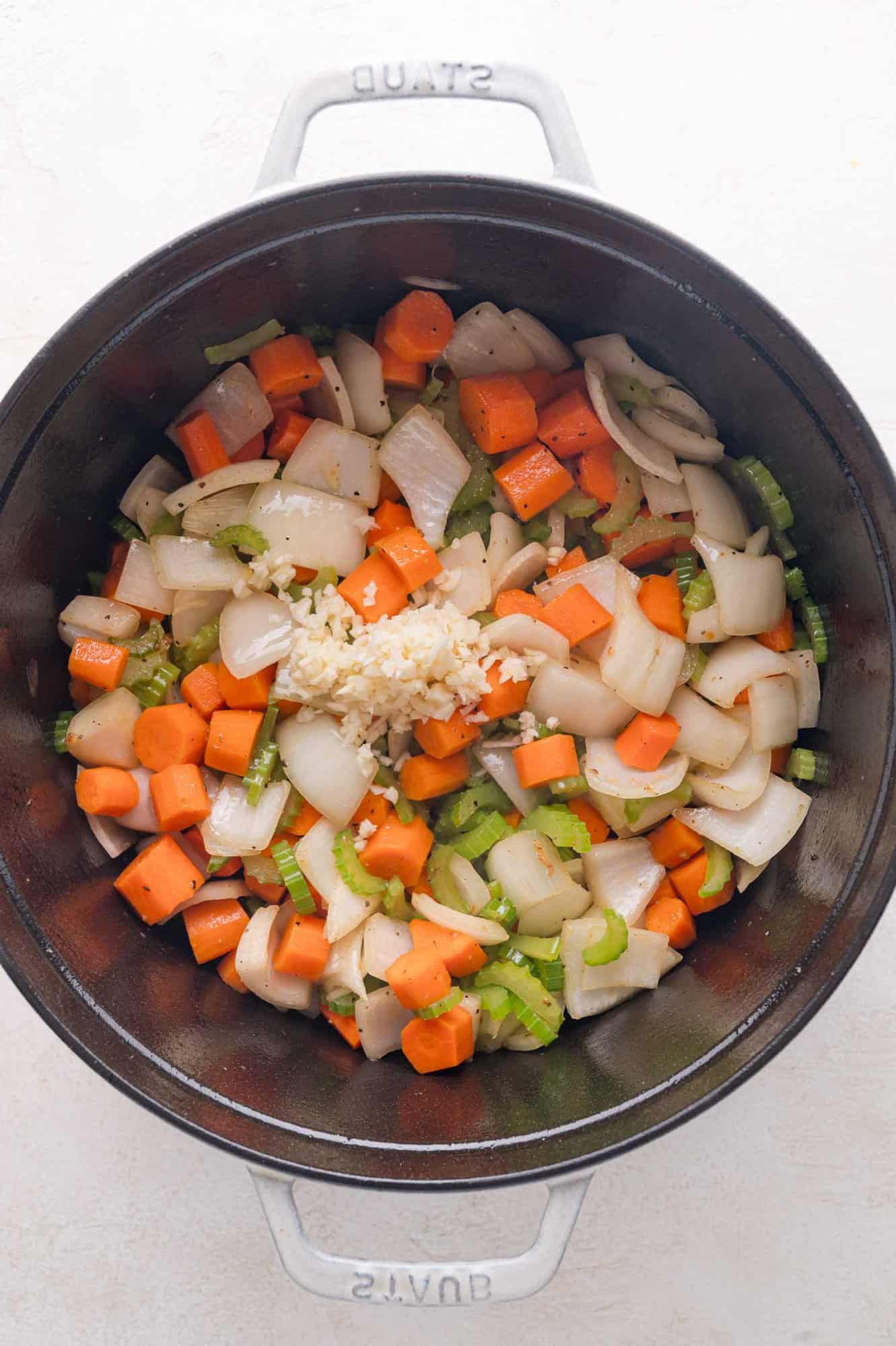 Chopped onions, carrots, and celery with minced garlic in a large black pot, viewed from above.