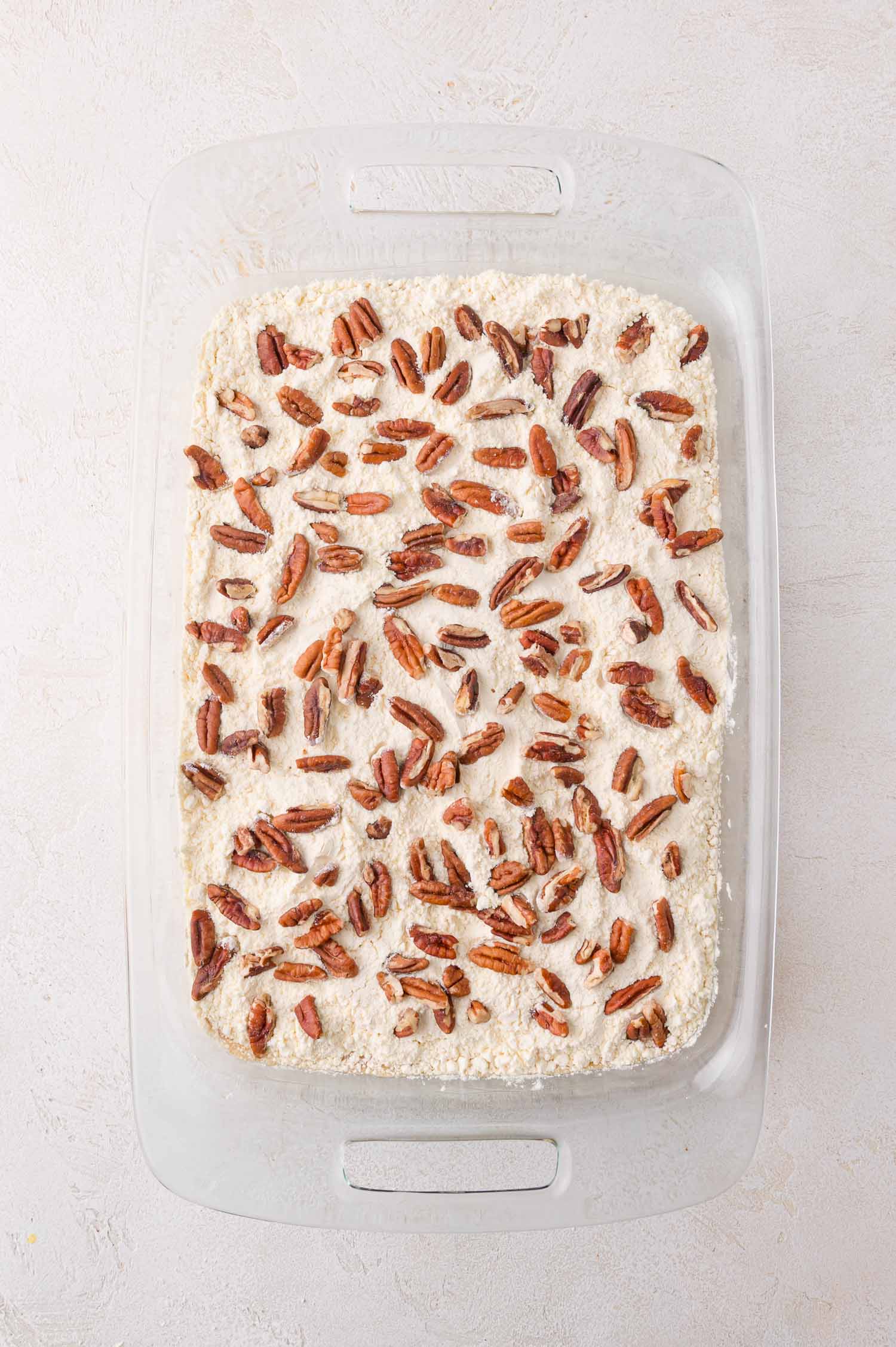 A glass baking dish filled with an unbaked pumpkin dump cake dessert topped with chopped pecans, set on a light-colored surface.