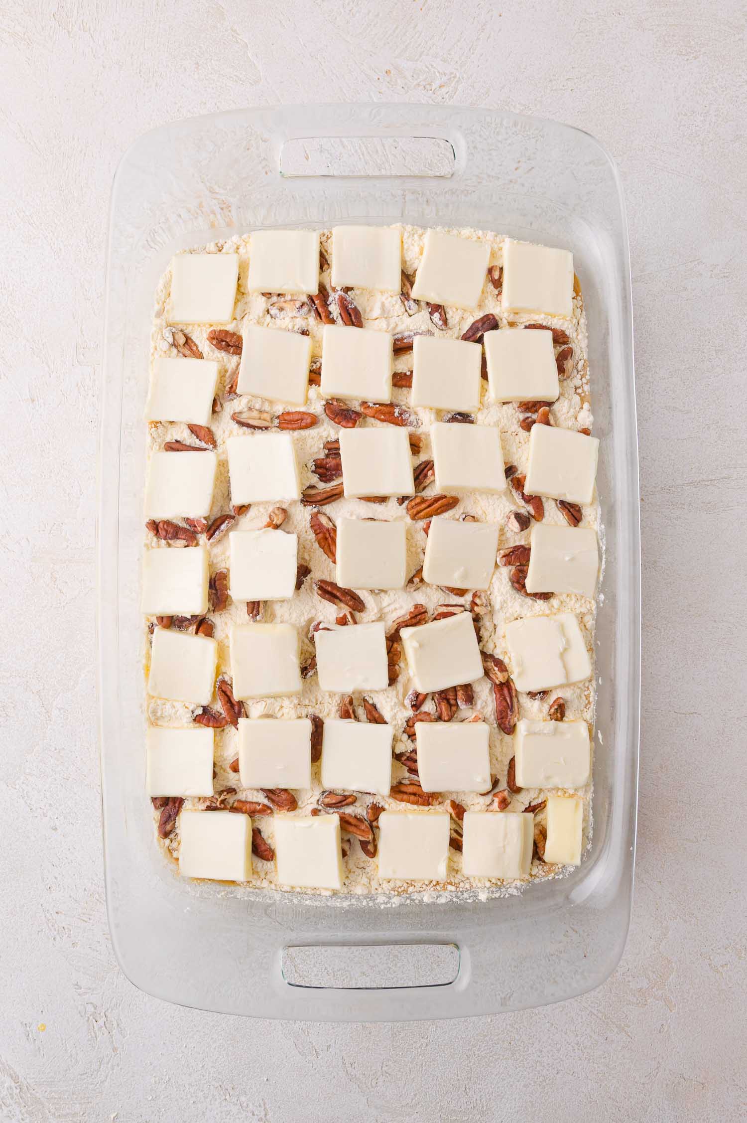 A glass baking dish filled with an unbaked dessert, topped with pecan halves and evenly spaced slices of butter on a crumbly mixture.