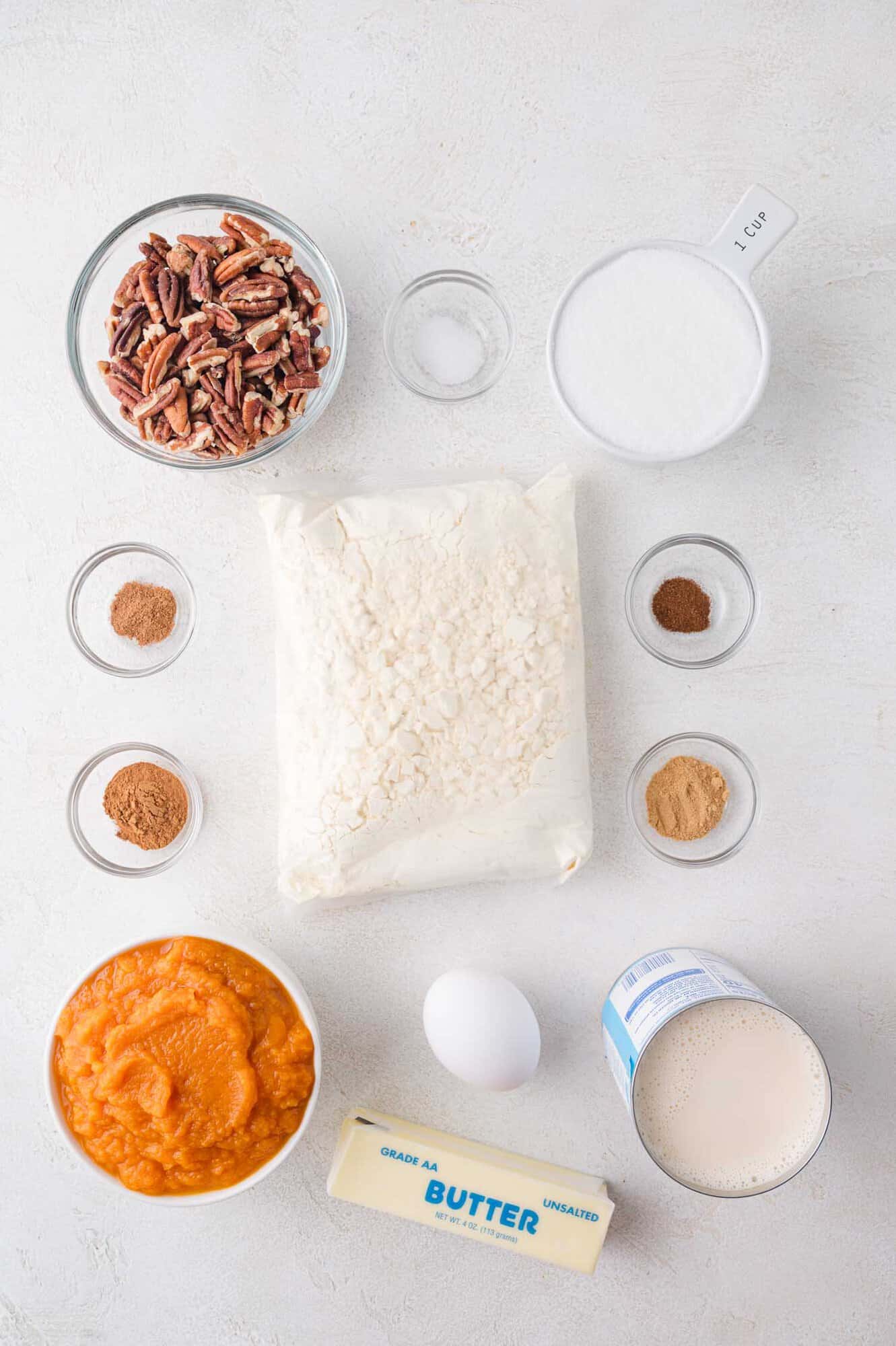 Ingredients for a recipe arranged on a white surface, including pecans, sugar, spices, flour, pumpkin puree, egg, butter, and evaporated milk.