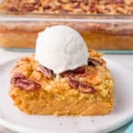 A square slice of pumpkin dump cake topped with pecans and a scoop of vanilla ice cream on a plate, with a baking dish of the dessert in the background.