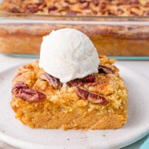 A square slice of pumpkin dump cake topped with pecans and a scoop of vanilla ice cream on a plate, with a baking dish of the dessert in the background.