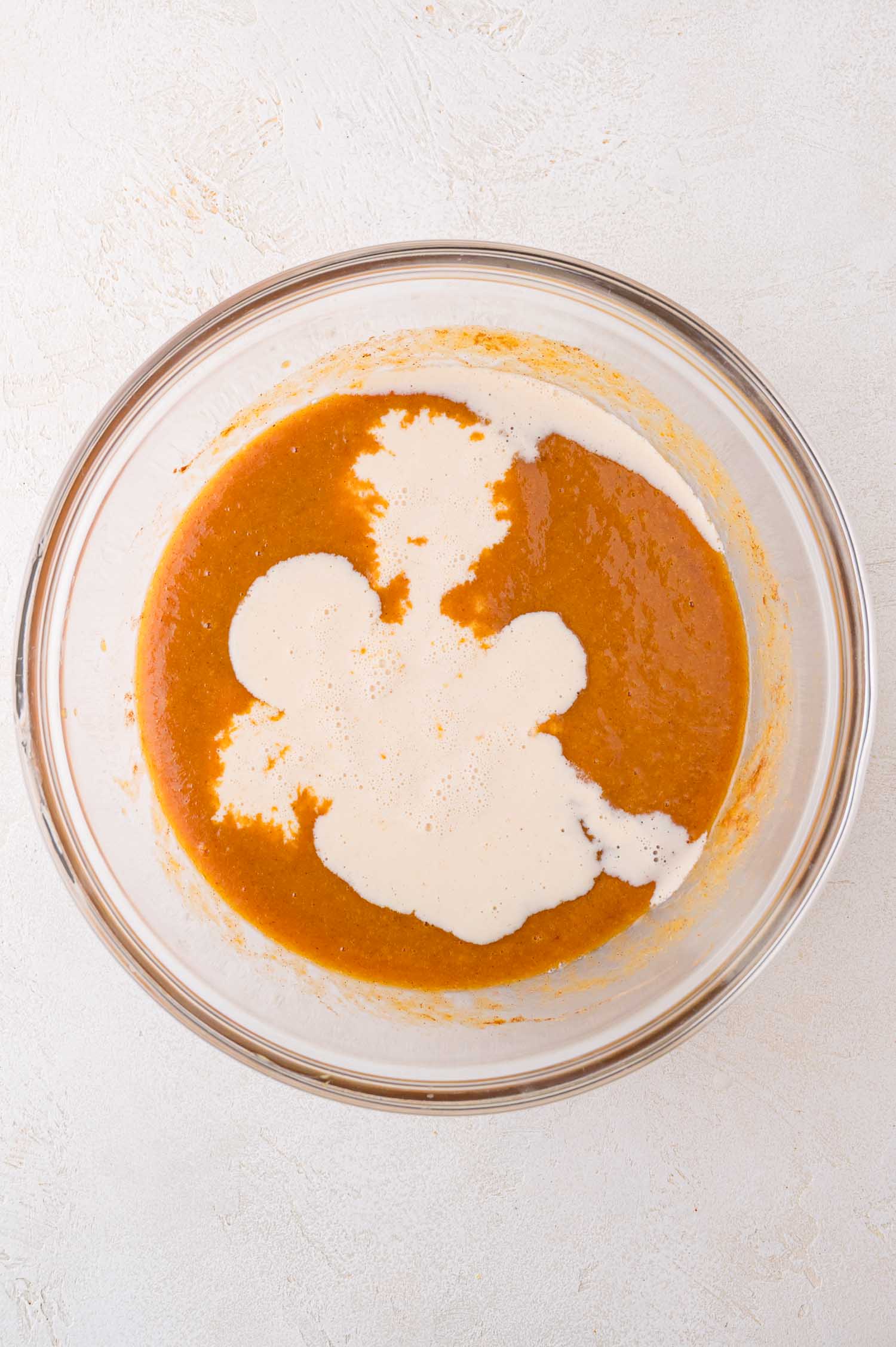 A glass bowl containing orange pumpkin puree with evaporated milk partially stirred in, sitting on a light-colored surface.