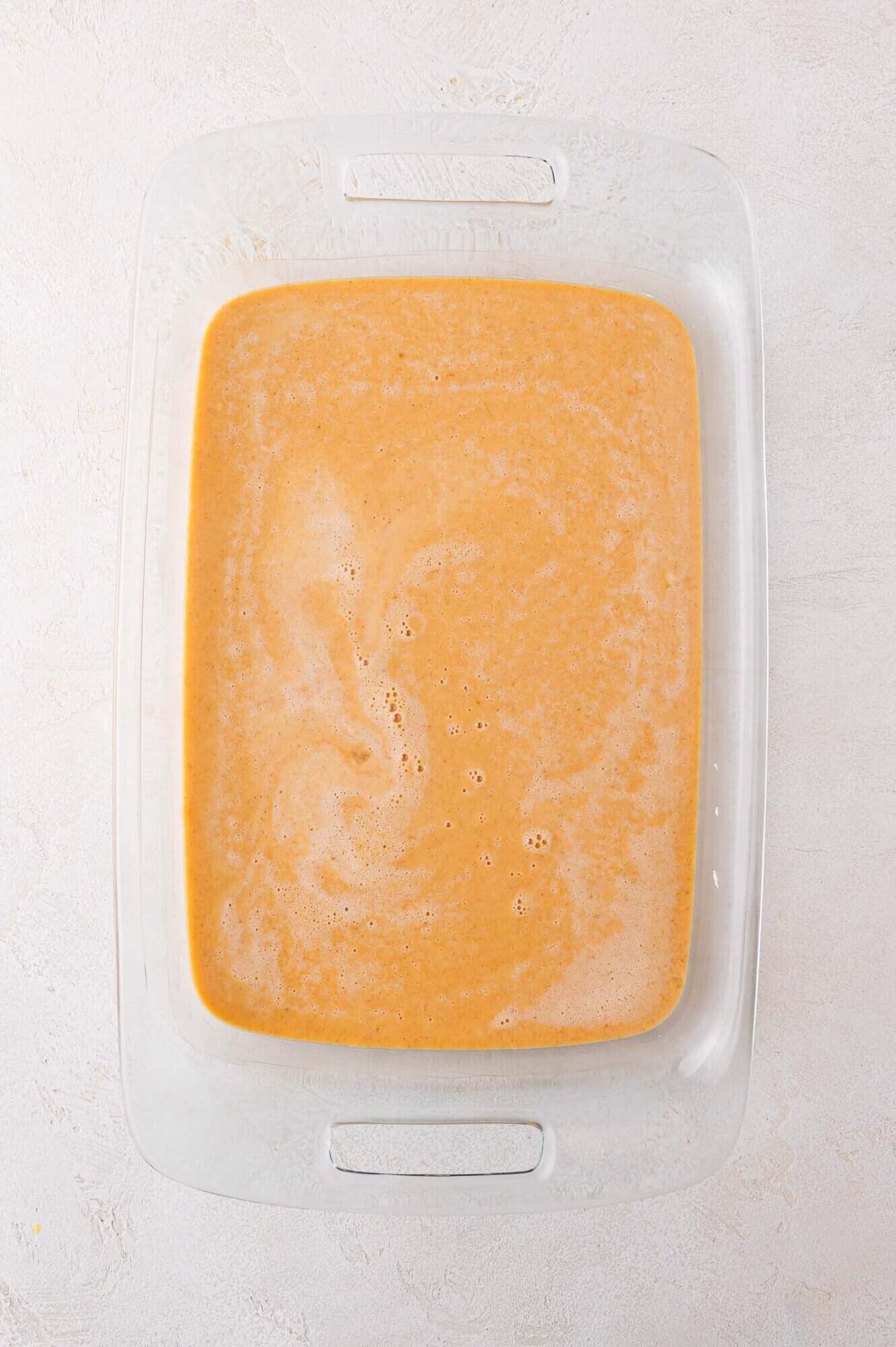 A clear glass baking dish filled with a smooth, orange-brown liquid (the pumpkin filling) mixture on a light textured surface.
