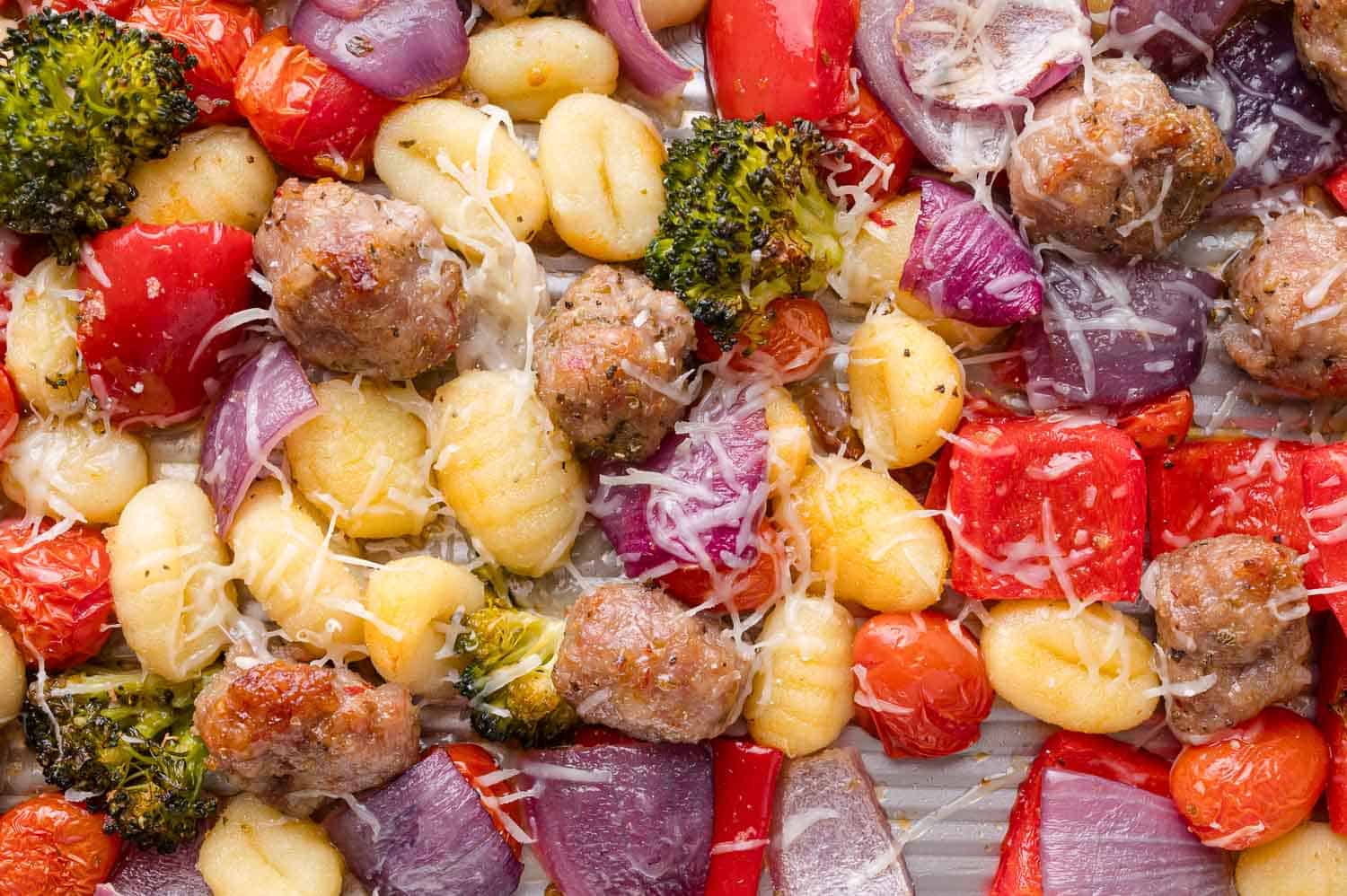 Sheet pan with cooked gnocchi, sausage, red onion, broccoli, red bell pepper, cherry tomatoes, and shredded cheese.