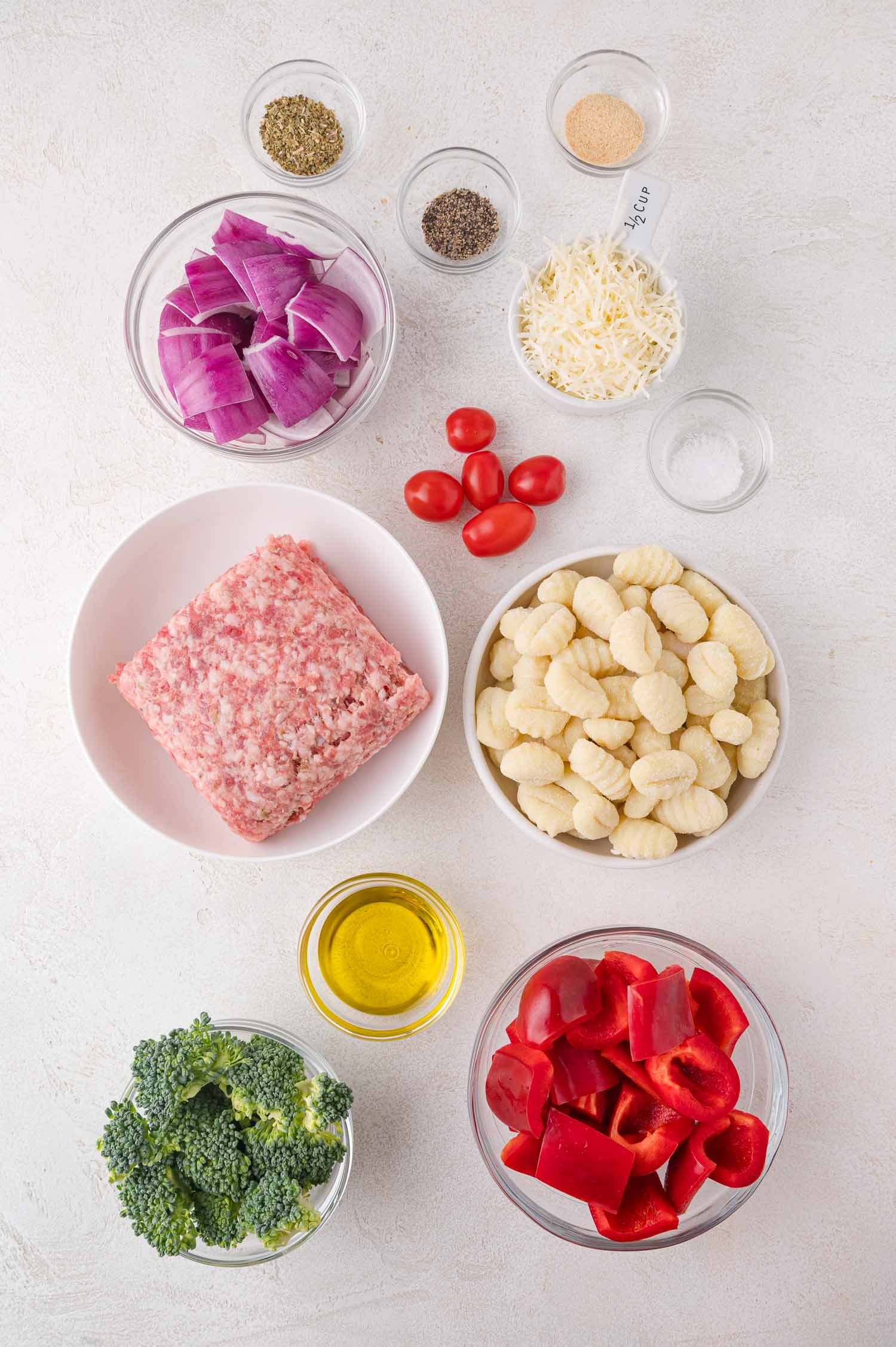 Assorted ingredients on a white surface, including ground sausage, gnocchi, broccoli, red bell pepper, cherry tomatoes, red onion, shredded cheese, spices, and olive oil in glass bowls.