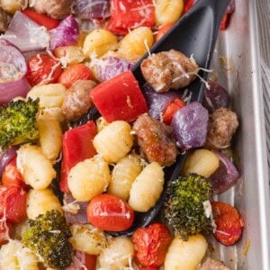 A sheet pan with cooked gnocchi, sausage, cherry tomatoes, red onion, red bell pepper, broccoli, and shredded cheese, with a black spatula scooping some of the food.