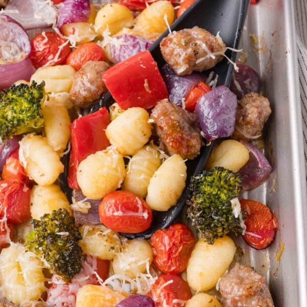 A tray of baked gnocchi with sausage, cherry tomatoes, broccoli, red onions, red bell peppers, and grated cheese, with a black serving spoon.
