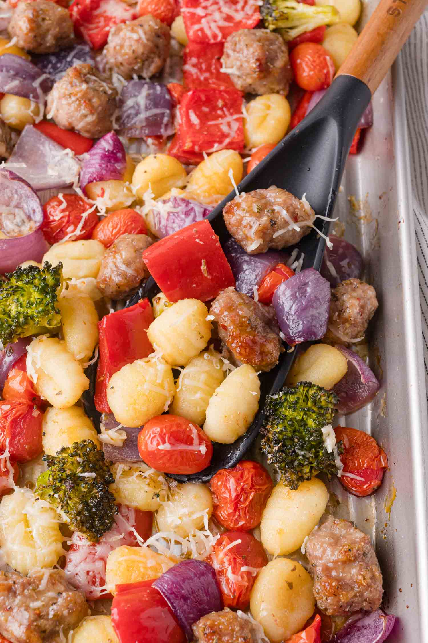 A sheet pan with cooked gnocchi, sausage, cherry tomatoes, red onion, red bell pepper, broccoli, and shredded cheese, with a black spatula scooping some of the food.