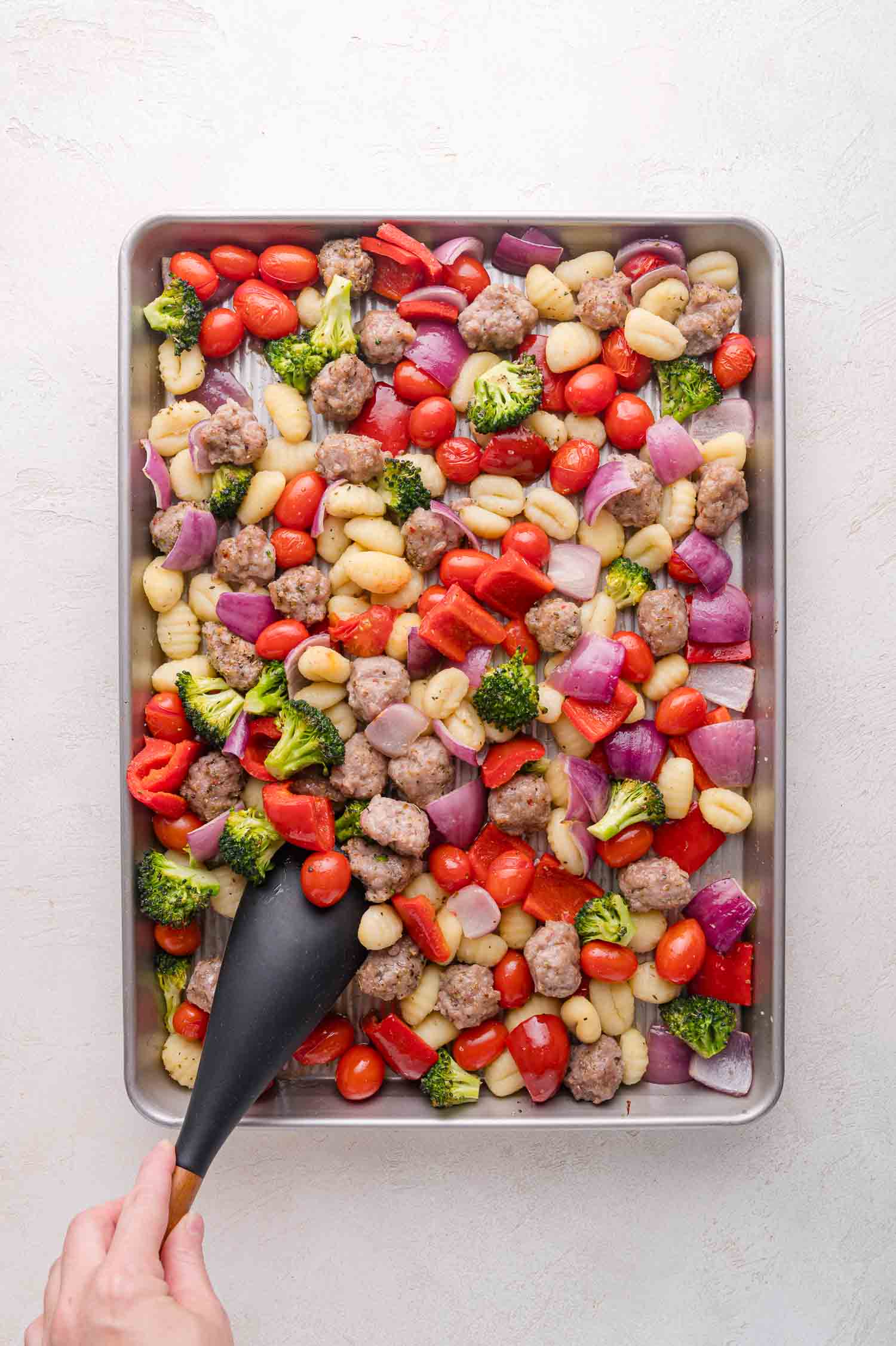 A sheet pan filled with gnocchi, cherry tomatoes, broccoli, red bell peppers, red onions, and sausage pieces, with a hand holding a black spatula.