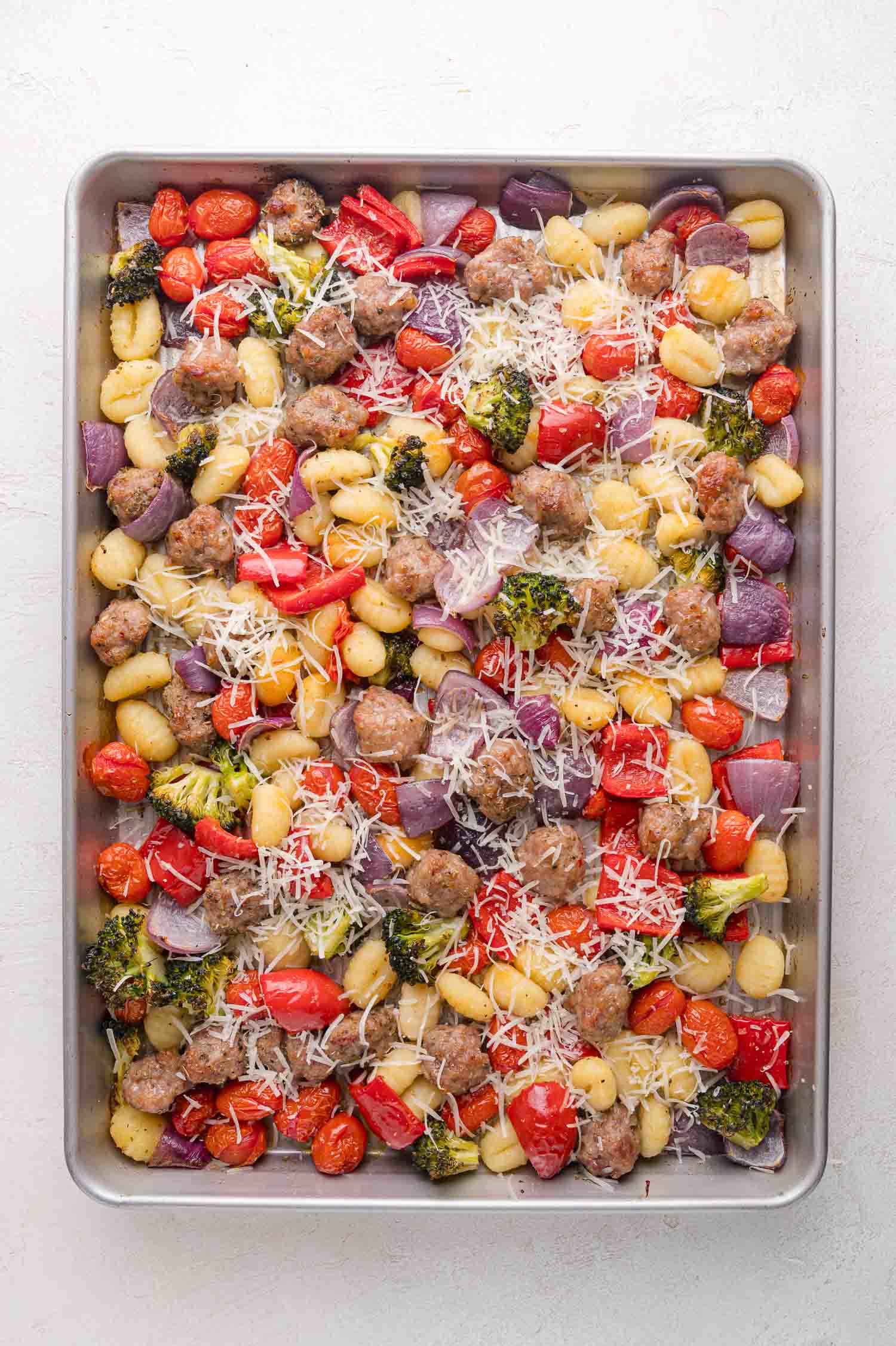 A sheet pan filled with cooked gnocchi, sausage, cherry tomatoes, red onion, broccoli, red bell peppers, and grated cheese.