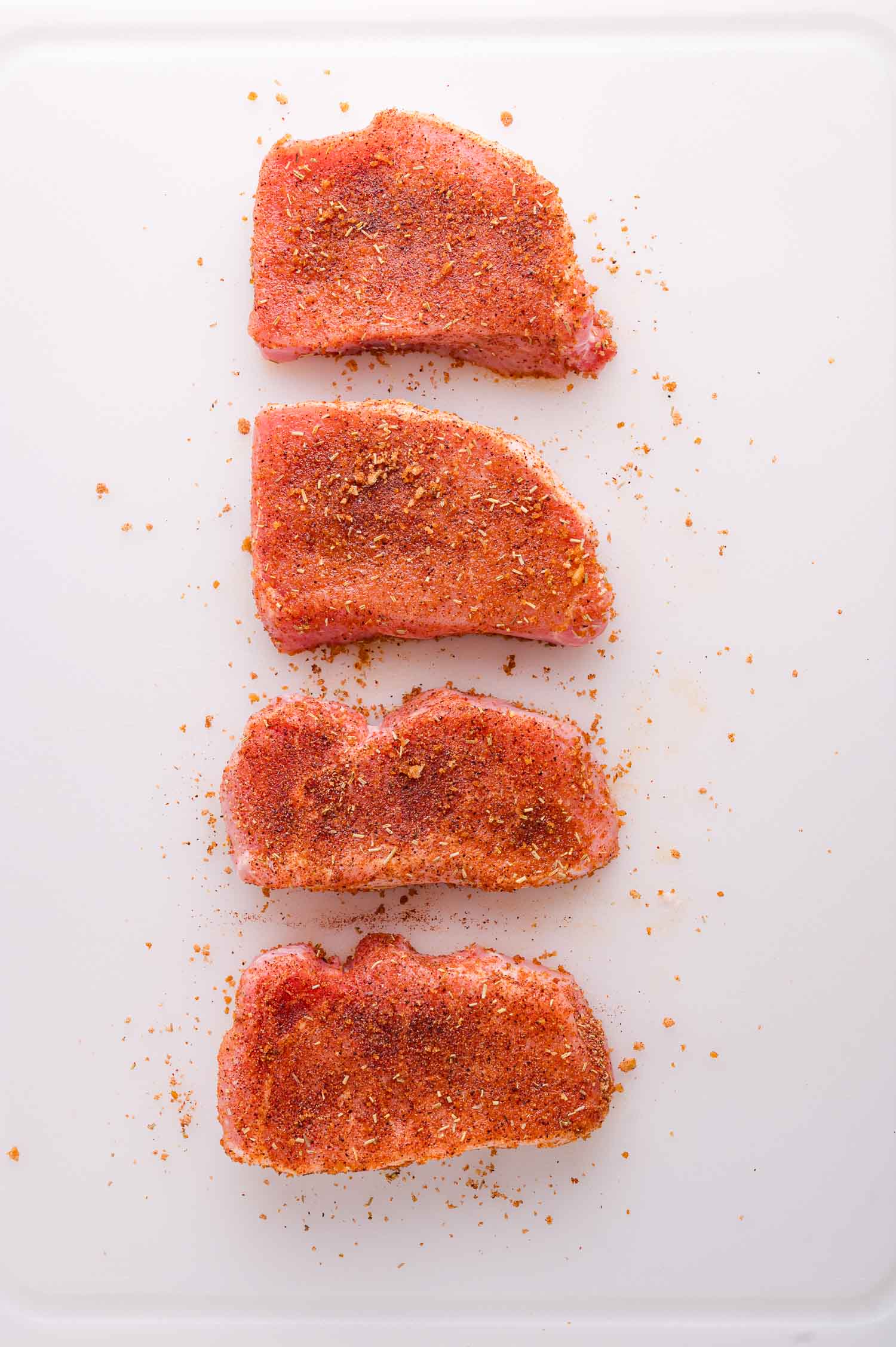Four raw pork chops coated with dry seasoning are arranged in a vertical line on a white surface, with some spices scattered around them.