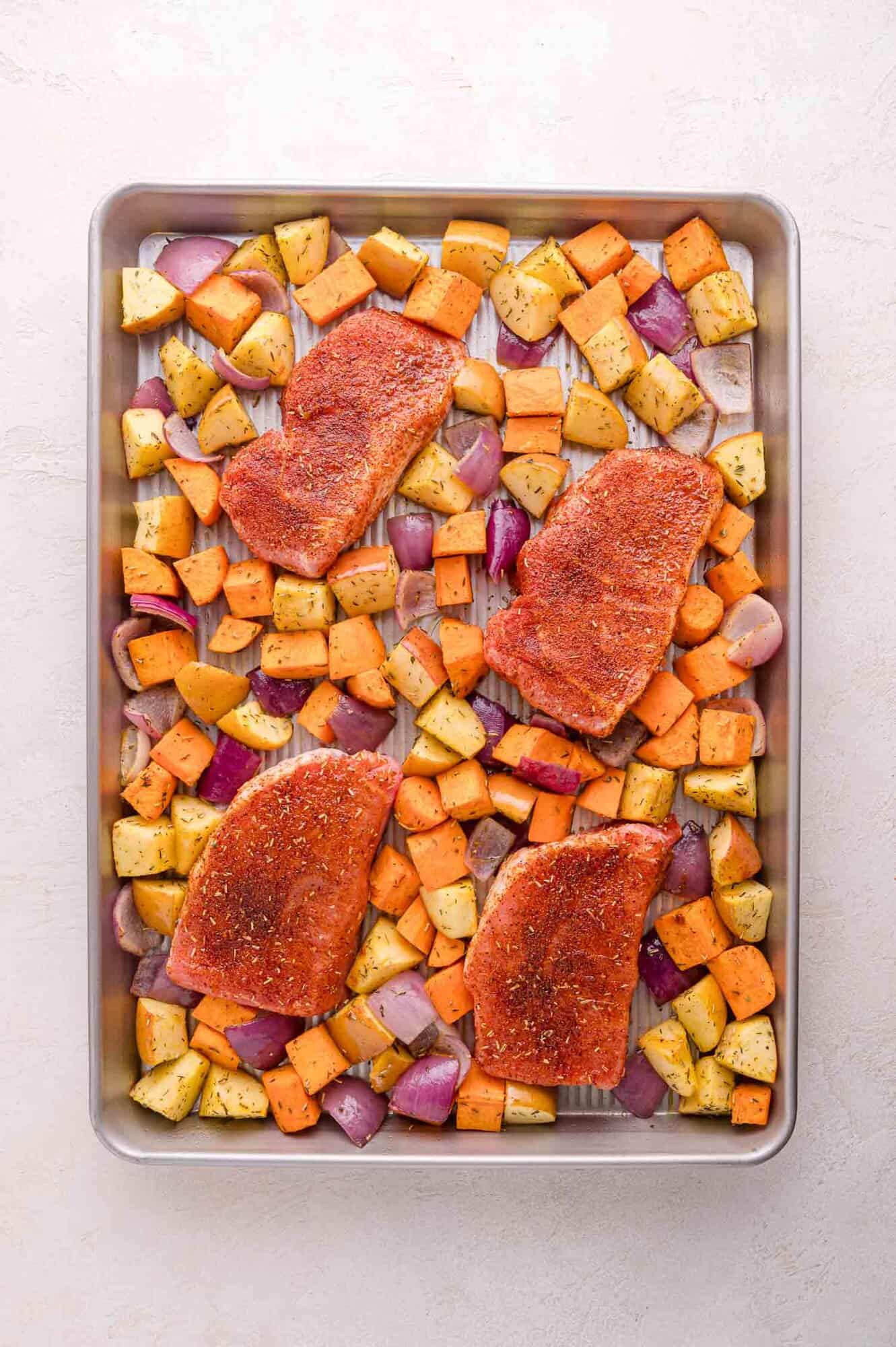 A sheet pan with four seasoned pork chops placed amongst diced sweet potatoes, apples, and red onions, ready to be cooked.