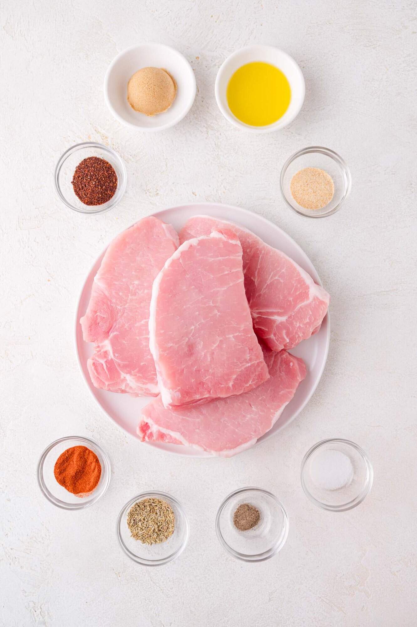 A plate of raw pork chops surrounded by small bowls of spices, seasoning, oil, and brown sugar on a white surface.